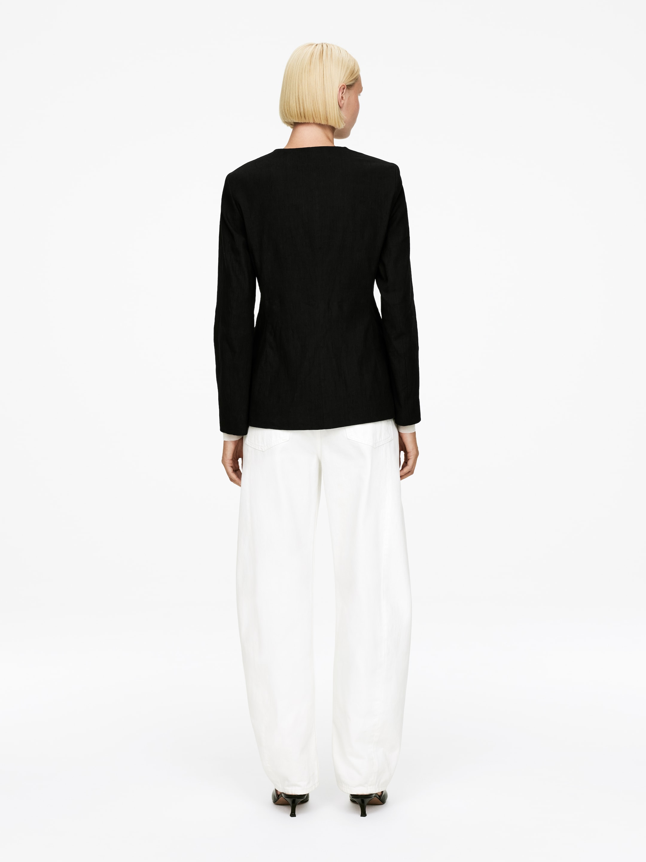 Ingrandisci l'immagine: Una giacca strutturata nera e pantaloni bianchi a gamba larga sono mostrati da dietro. La giacca presenta uno scollo senza colletto e maniche lunghe, con un indumento bianco che si stratifica sotto i polsini. I pantaloni bianchi mostrano una silhouette a gamba a palloncino, raccogliendosi alle caviglie.