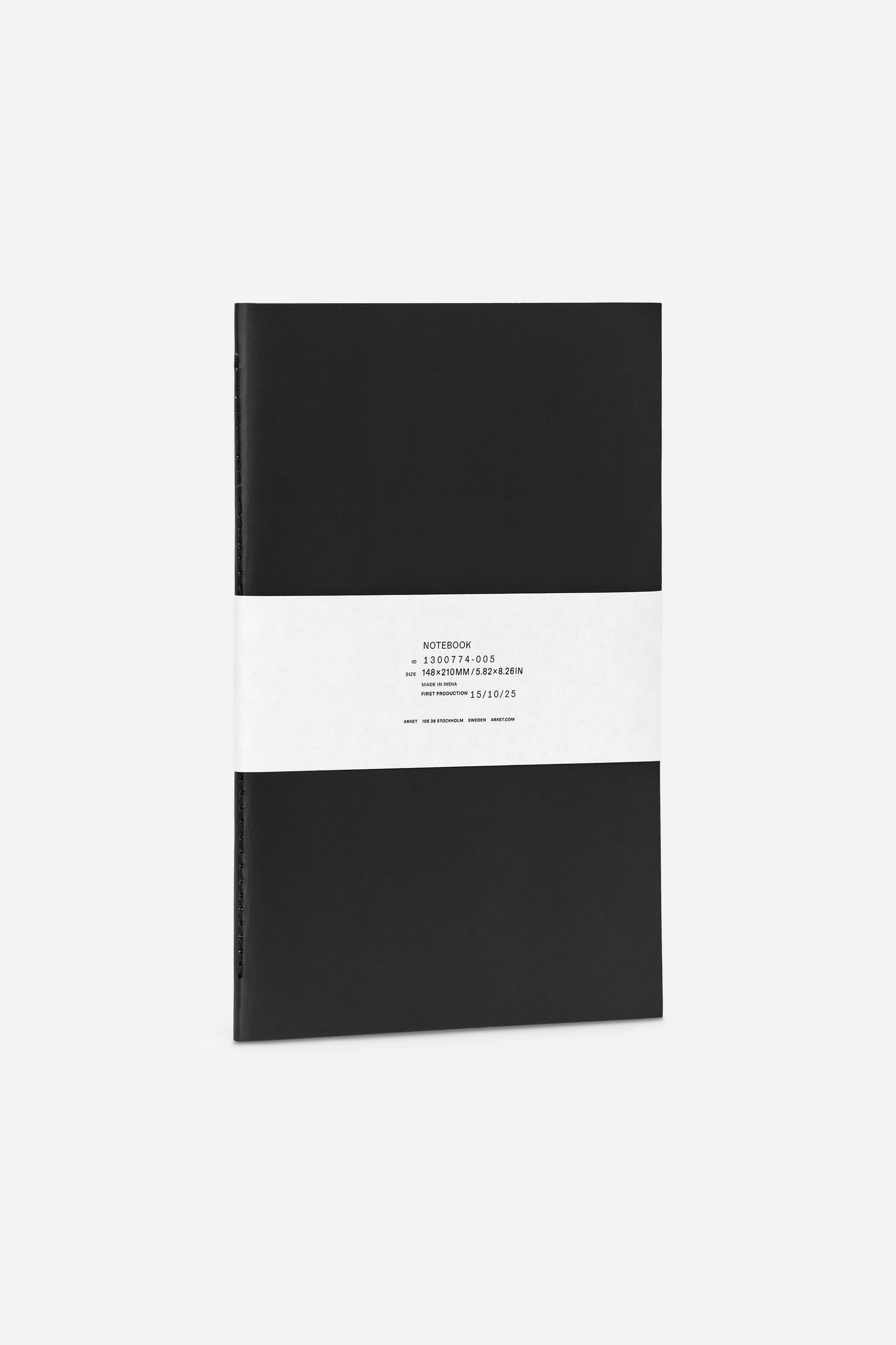 Größeres Bild anzeigen: Ein A5-Notizbuch präsentiert sein mattes schwarzes Cover, das nach vorne zeigt. Ein weißes Papierband umwickelt das Notizbuch horizontal und zeigt gedruckten Text mit Produktspezifikationen und Produktionsdetails. Eine genähte Bindung ist entlang der linken Kante des Notizbuchs sichtbar.