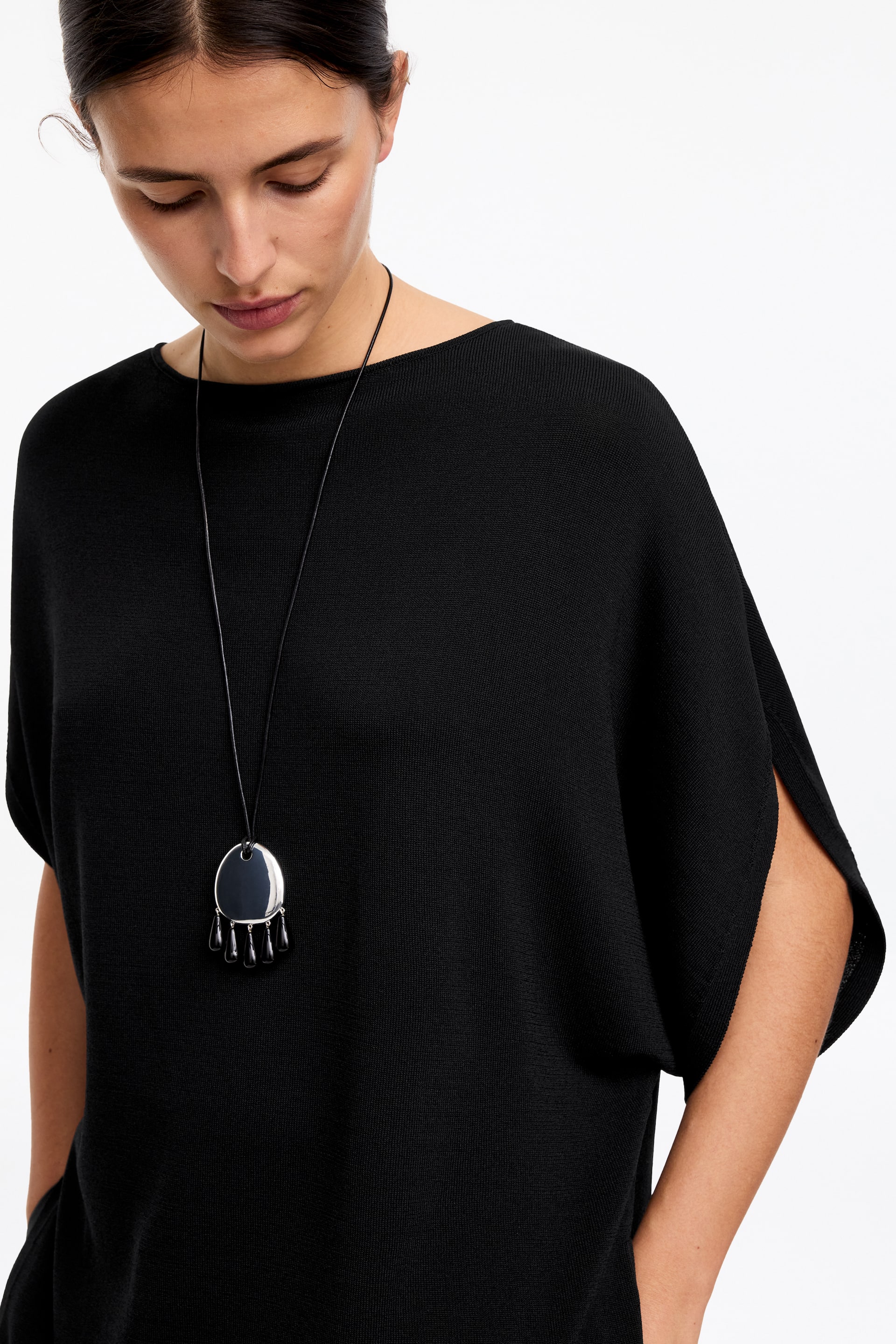 View larger image: A black knitted top features a boat neck and wide dolman sleeves with open slits along the outer arm. An oval, silver-toned pendant necklace adorned with dark teardrop elements is layered over the front of the top.