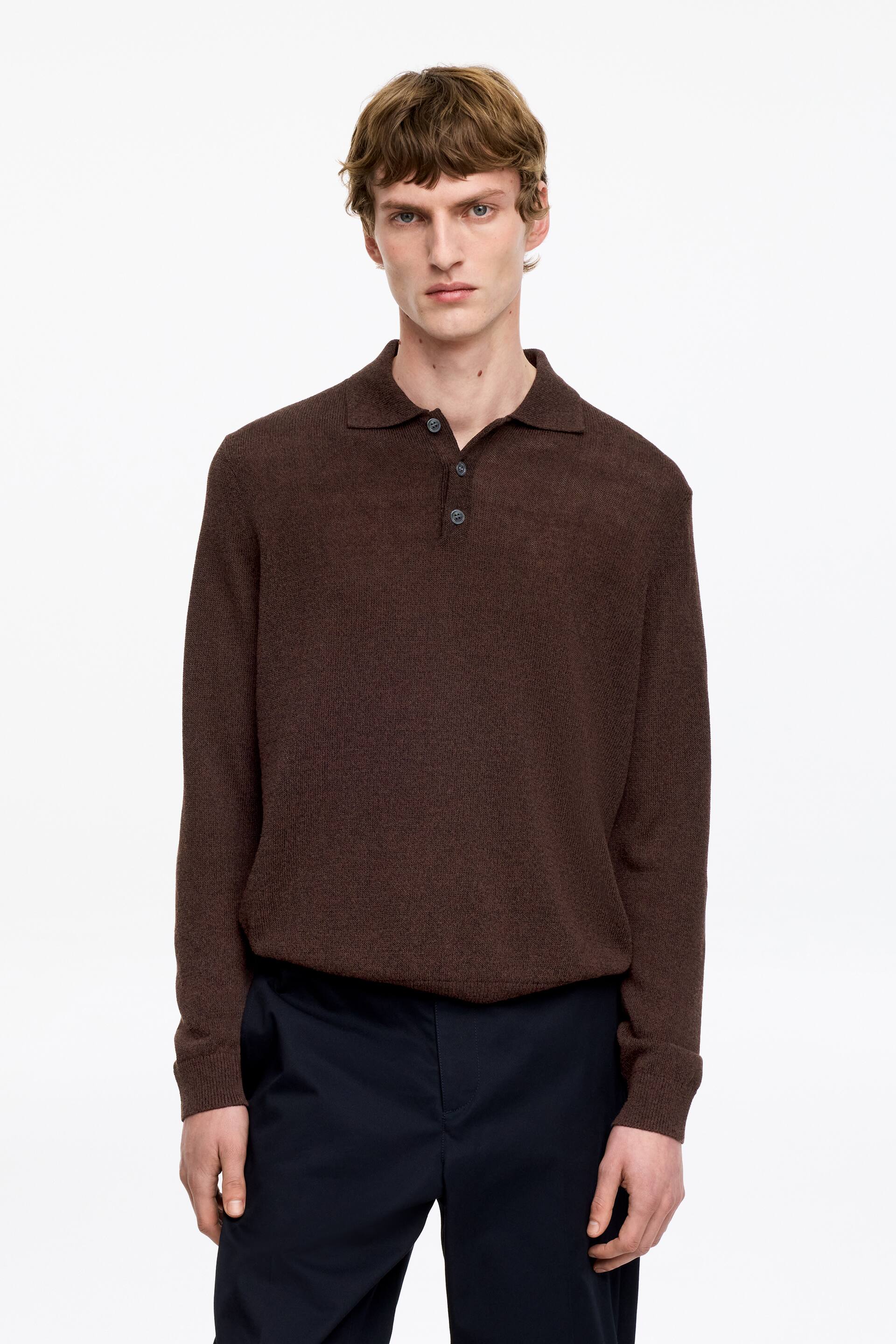 Ver imagem maior: Uma camisola de malha castanha de manga comprida apresenta uma gola polo e uma carcela de três botões com dois botões abotoados. Exibe mangas compridas com punhos canelados e um corte descontraído, atualmente por dentro das calças.