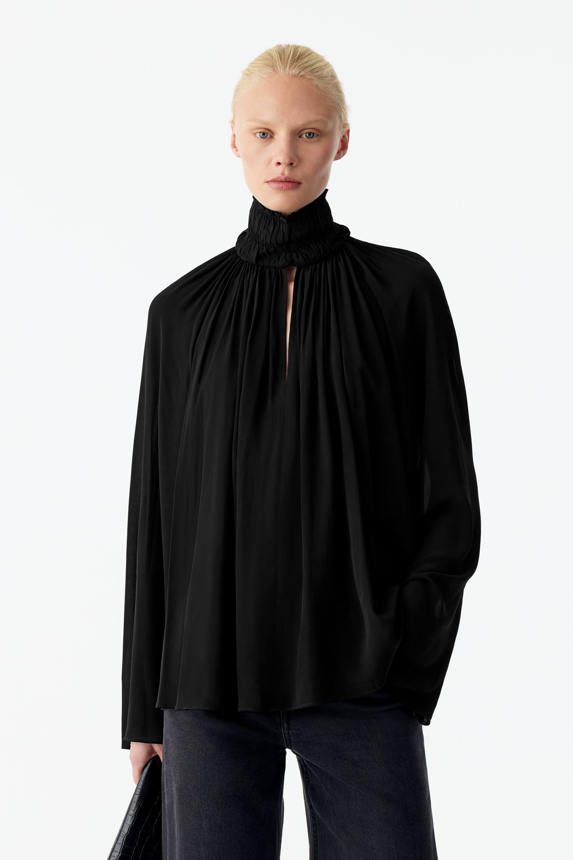 Visualizza l'immagine ingrandita: La modella indossa una blusa nera a maniche lunghe, che mette in risalto il suo collo alto con smock, la fessura frontale e il tessuto fluido arricciato che crea una silhouette voluminosa. La blusa è abbinata a jeans grigio scuro, mentre la modella tiene in mano una borsa nera testurizzata.