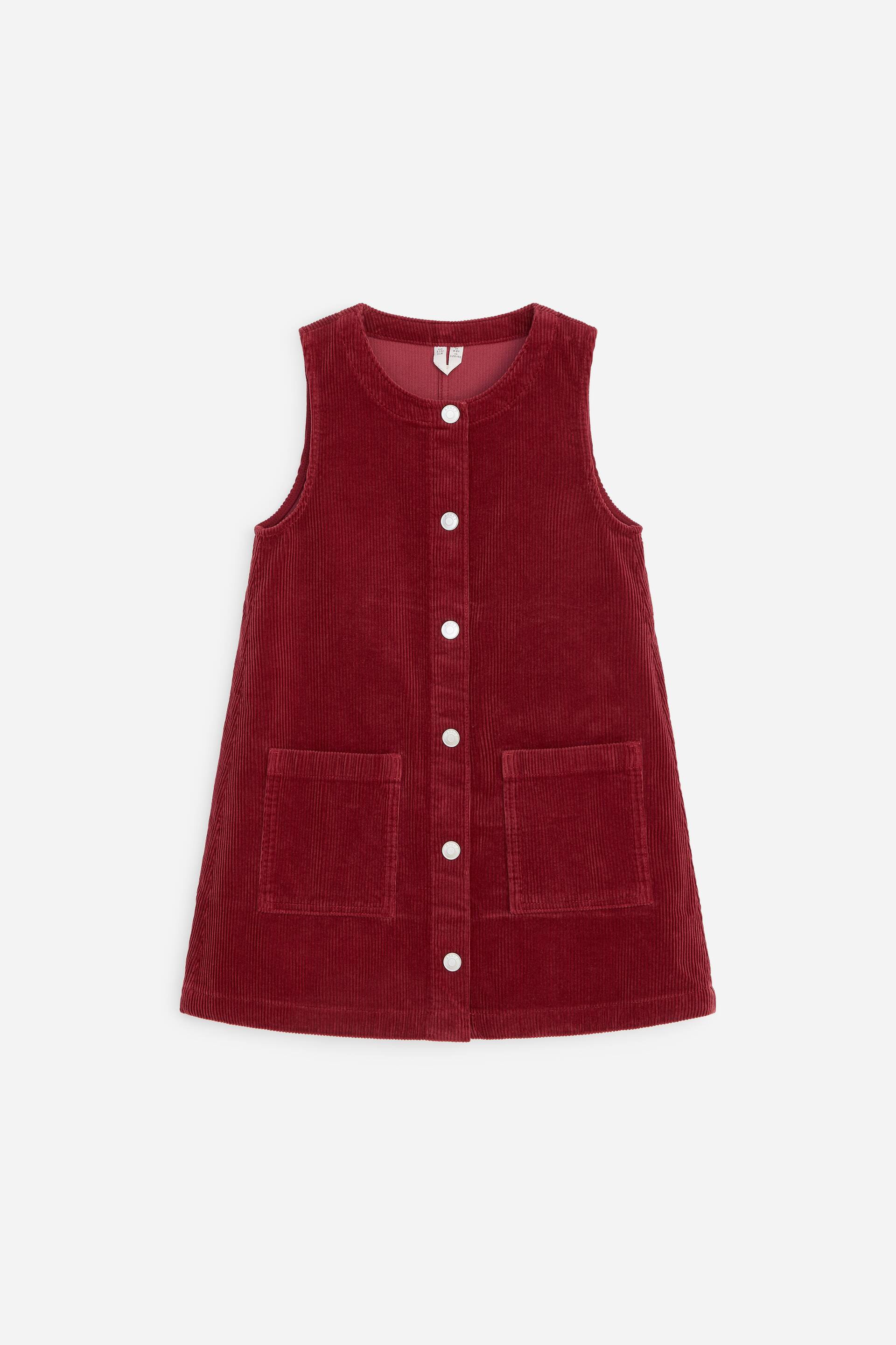 Ver imagen más grande: Un vestido tipo peto de pana roja se muestra de frente, con un escote redondeado, una tapeta frontal de botones de largo completo con botones blancos y dos grandes bolsillos de parche en la parte delantera.