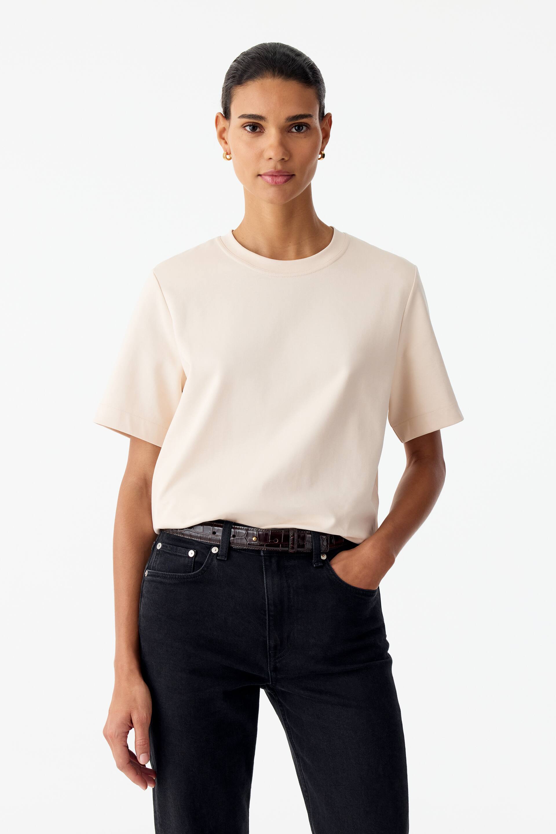 Agrandir l'image: Un T-shirt en coton de couleur crème, doté d’un col rond classique et de manches courtes structurées, est porté par le modèle. Le T-shirt est rentré dans un jean noir, accompagné d’une ceinture marron foncé.