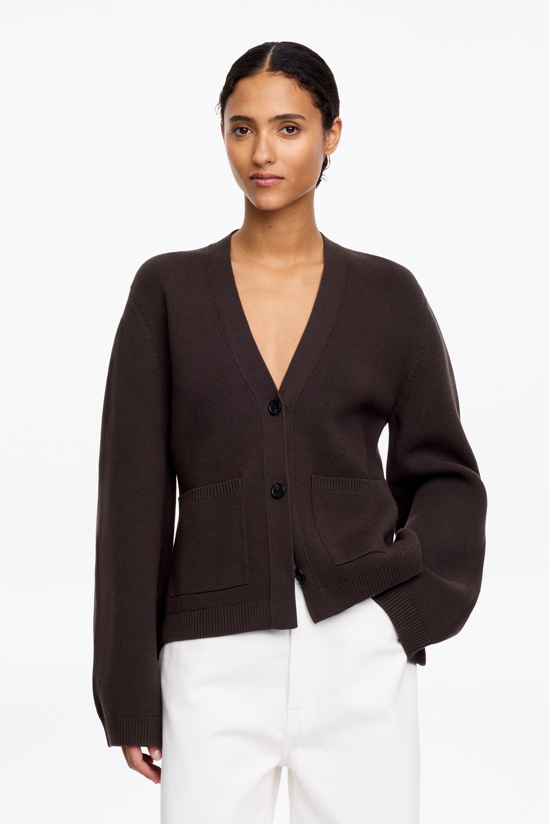 Vis større bilde: En mørkebrun cardigan med V-hals, tre sorte knapper foran hvor den øverste knappen er festet, og to påsydde lommer nær kanten. Den har lange ermer med ribbestrikkede mansjetter og en ribbestrikket kant nederst, vendt fremover.