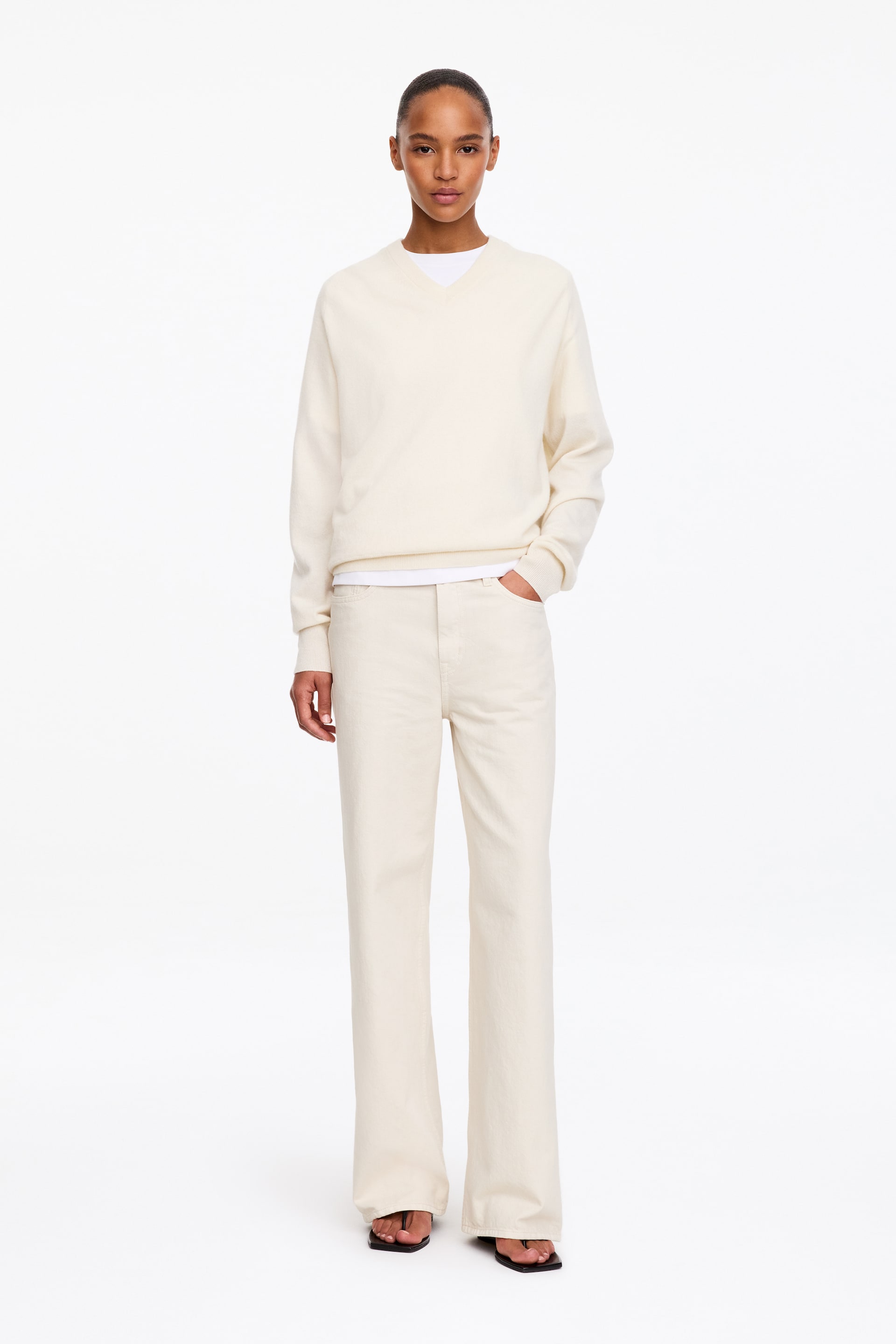 Agrandir l'image: Un jean en denim blanc cassé, taille haute, à la coupe décontractée et aux jambes larges, porté par le mannequin, qui se tient avec une main dans la poche avant. Le jean est associé à un pull crème à col en V, porté par-dessus un t-shirt blanc.
