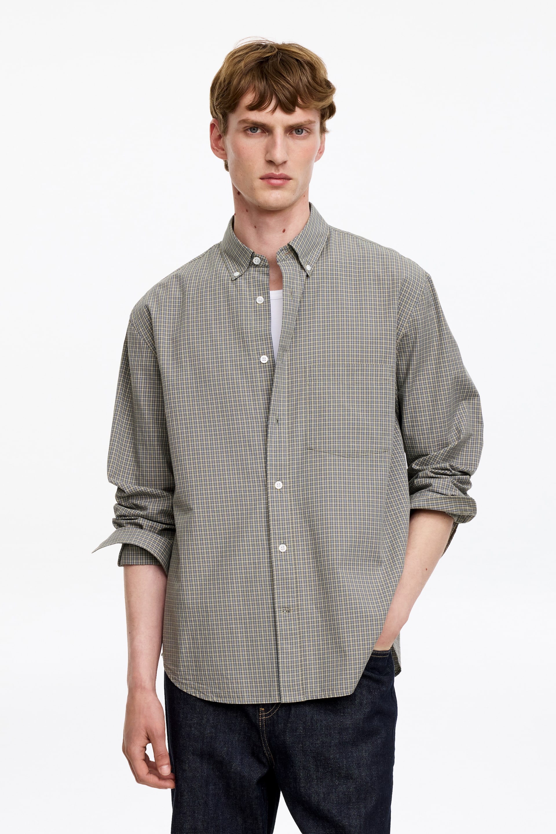 Agrandir l'image: Une chemise en coton avec un motif à carreaux teint en fil, un col boutonné, une poche poitrine unique et un ourlet arrondi est portée par le mannequin. La chemise à coupe décontractée est stylée partiellement déboutonnée sur un sous-vêtement blanc, avec l'un de ses poignets boutonnés retroussé, et le mannequin se tient avec une main dans la poche d'un jean en denim foncé.