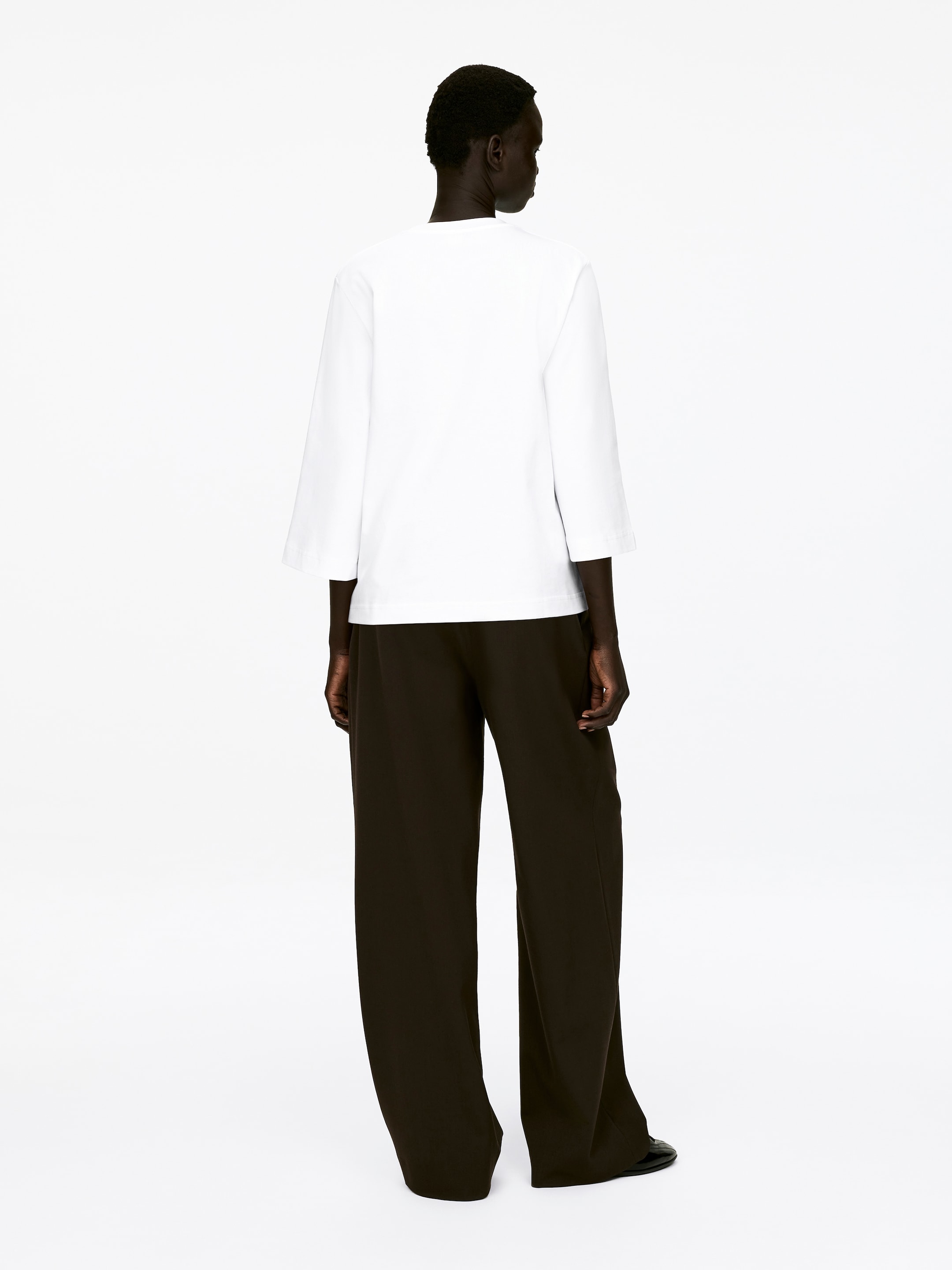 Agrandir l'image: Un T-shirt blanc à col ras du cou, confectionné dans un jersey de coton épais et frais et doté de manches 3/4, est porté par le mannequin. Le T-shirt est assorti à un pantalon large de couleur sombre.