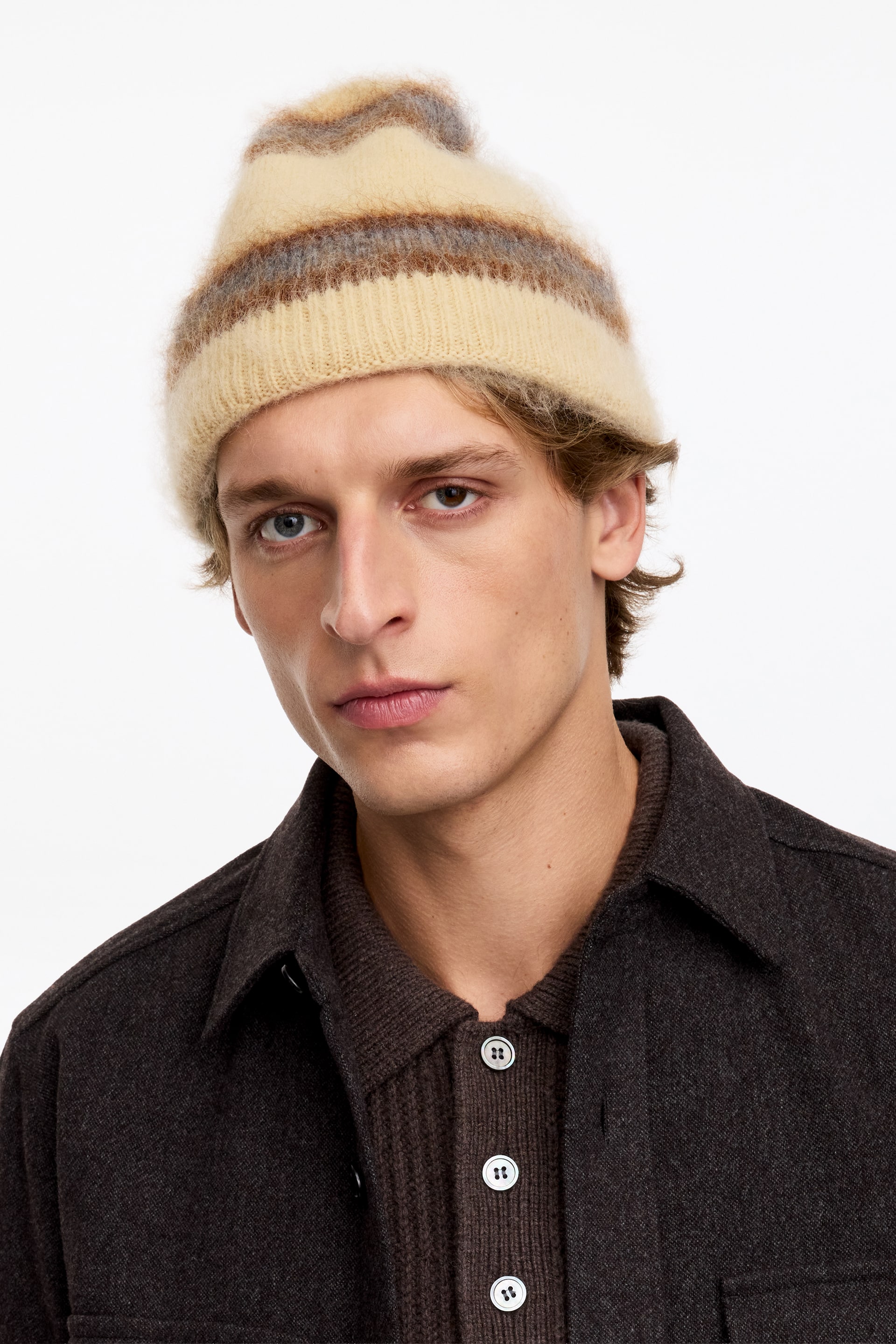 Zobacz większy obraz: Model ma na sobie miękką, teksturowaną czapkę typu beanie z mieszanki moheru, ozdobioną poziomymi paskami w kolorze kremowym, brązowym i szarym. Czapka ma prążkowany, wywijany brzeg i została zestawiona z ciemnobrązową koszulową kurtką z kołnierzem.