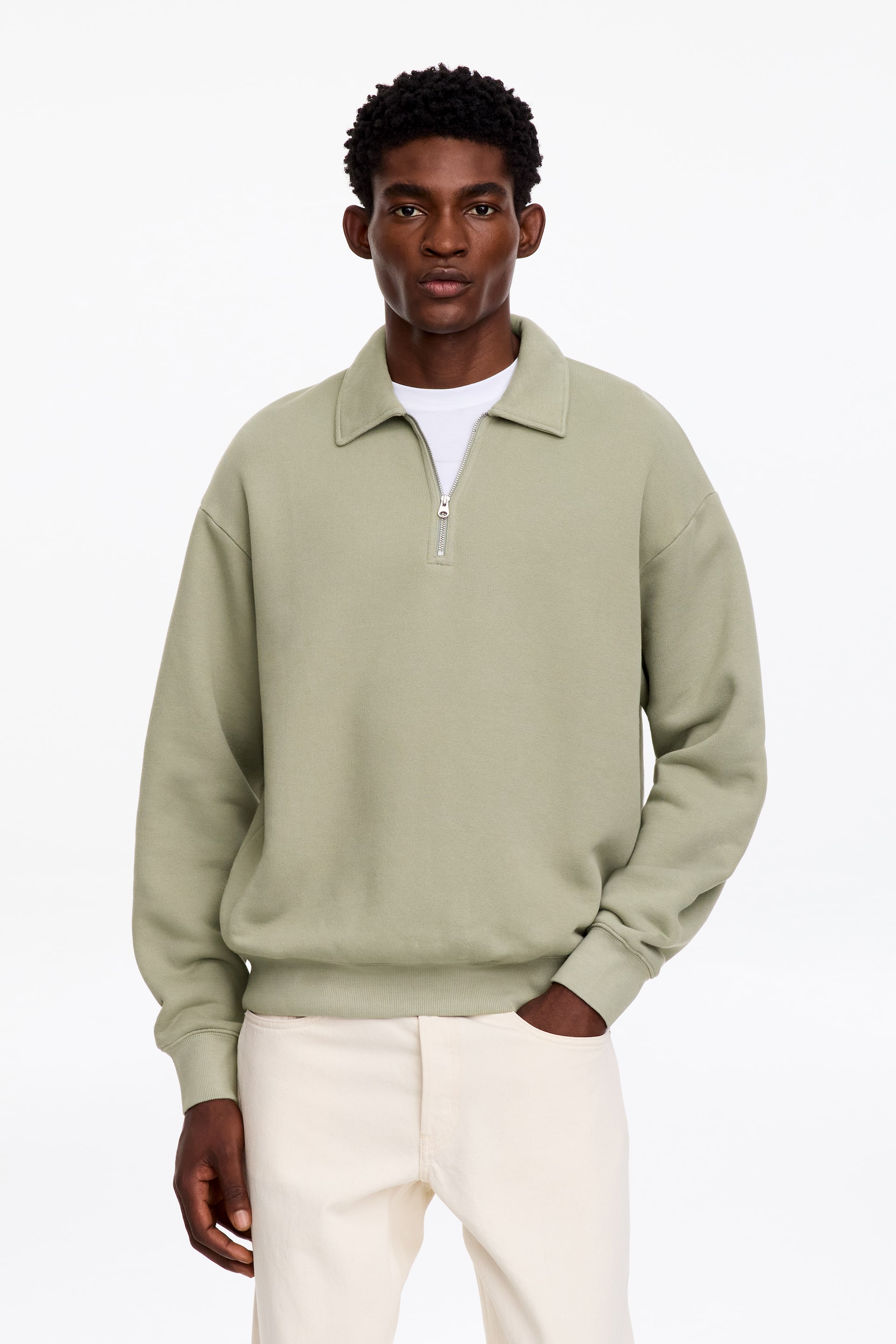 Grösseres Bild ansehen: Ein hellgrüner Sweatshirt mit Kragen und silbernem halbem Reissverschluss wird vom Model über einem weissen T-Shirt getragen. Der Sweatshirt verfügt über gerippte Bündchen und einen gerippten Saum und wird mit cremeweissen Hosen kombiniert.