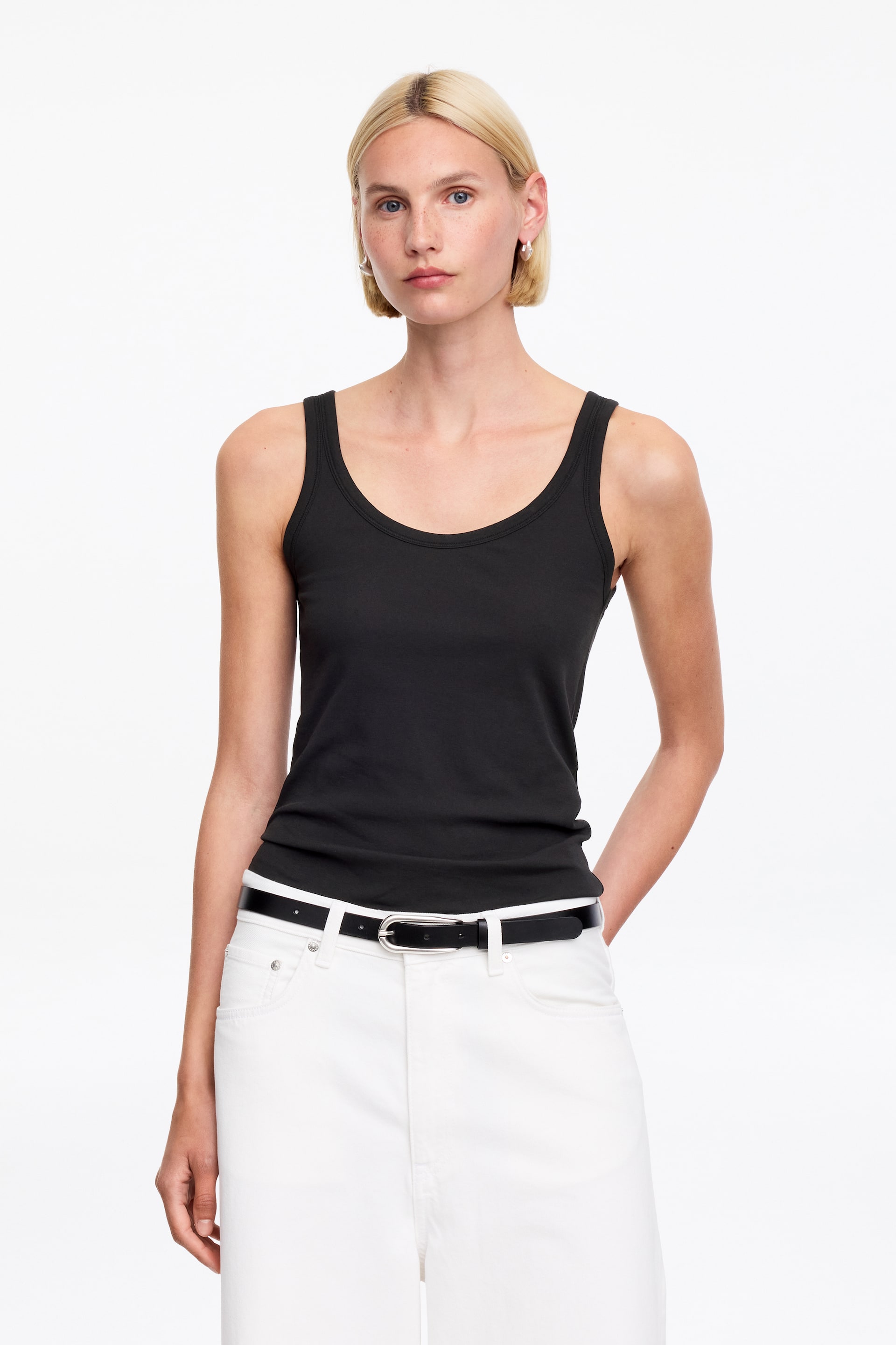 Vis større bilde: En svart tanktopp med rund hals og brede skulderstropper. Den har en tettsittende silhuett og er stukket inn i hvite bukser.