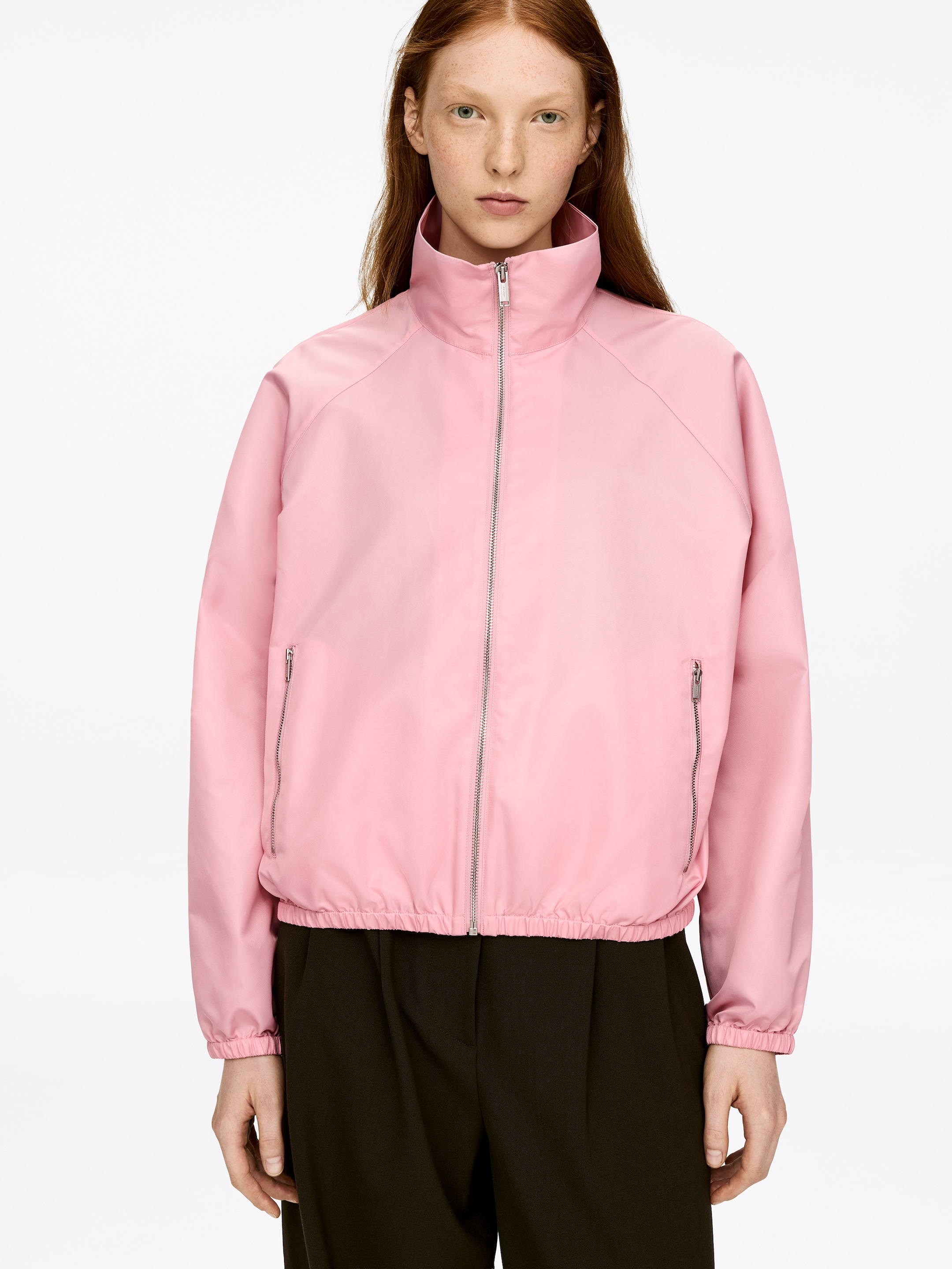 Vis større bilde: En rosa sporty jakke i nylon bæres av modellen. Den har en høy krage, en sentral metallglidelås, to metallglidelåslommer, og elastikk nederst på kanten og på mansjettene.