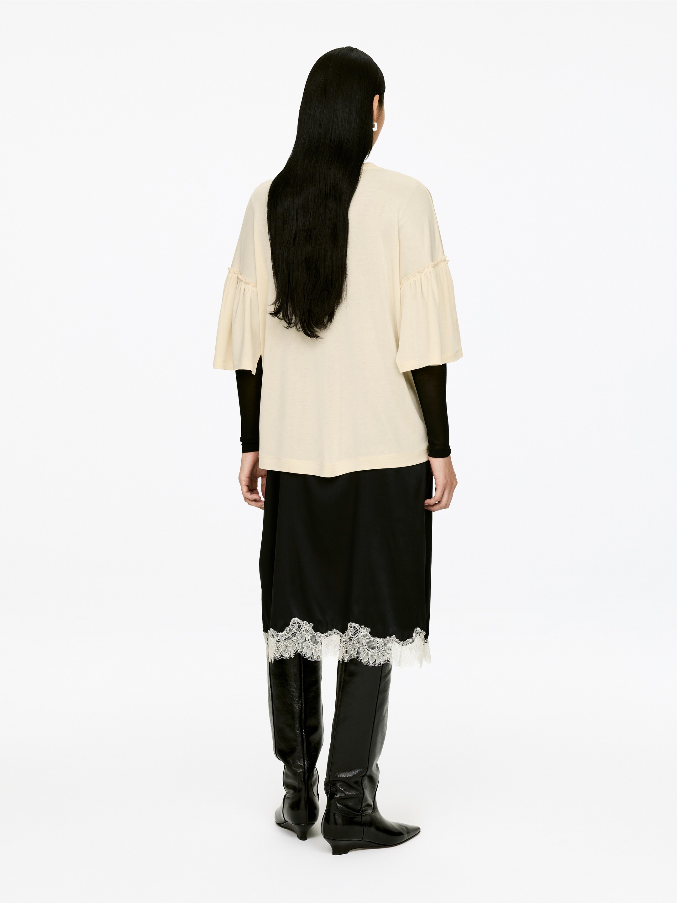 Visa större bild: En krämfärgad T-shirt med rund hals, med fransiga ärmar och en vanlig passform, bärs av modellen. Den är lagd över en svart långärmad topp och en svart kjol med spetskant.