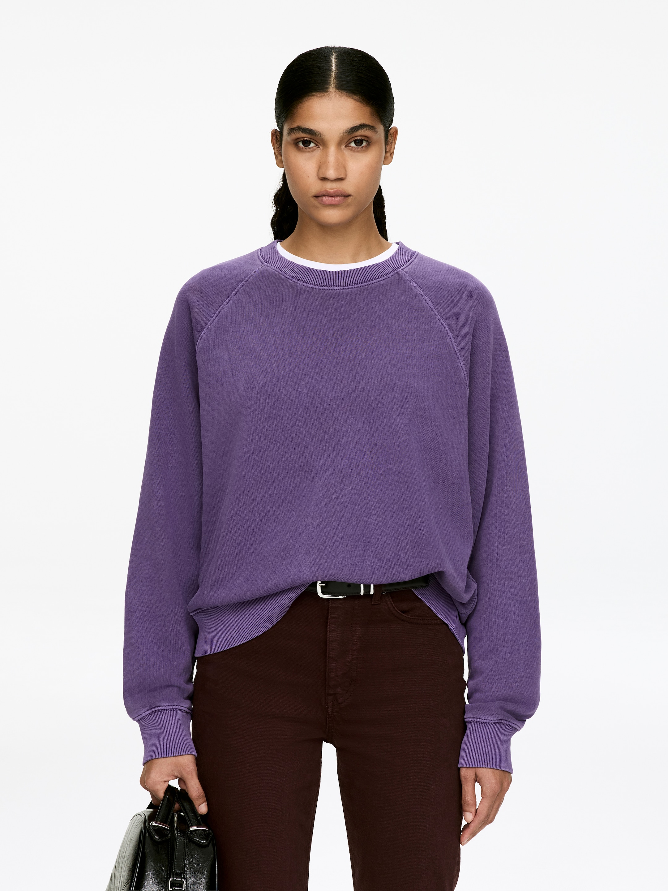 Grösseres Bild ansehen: Ein violettes Sweatshirt mit Rundhalsausschnitt, das eine normale Passform, Raglanärmel und gerippte Abschlüsse aufweist, wird vom Model getragen. Ein weißes Oberteil ist unterhalb des Halsausschnitts zu sehen.