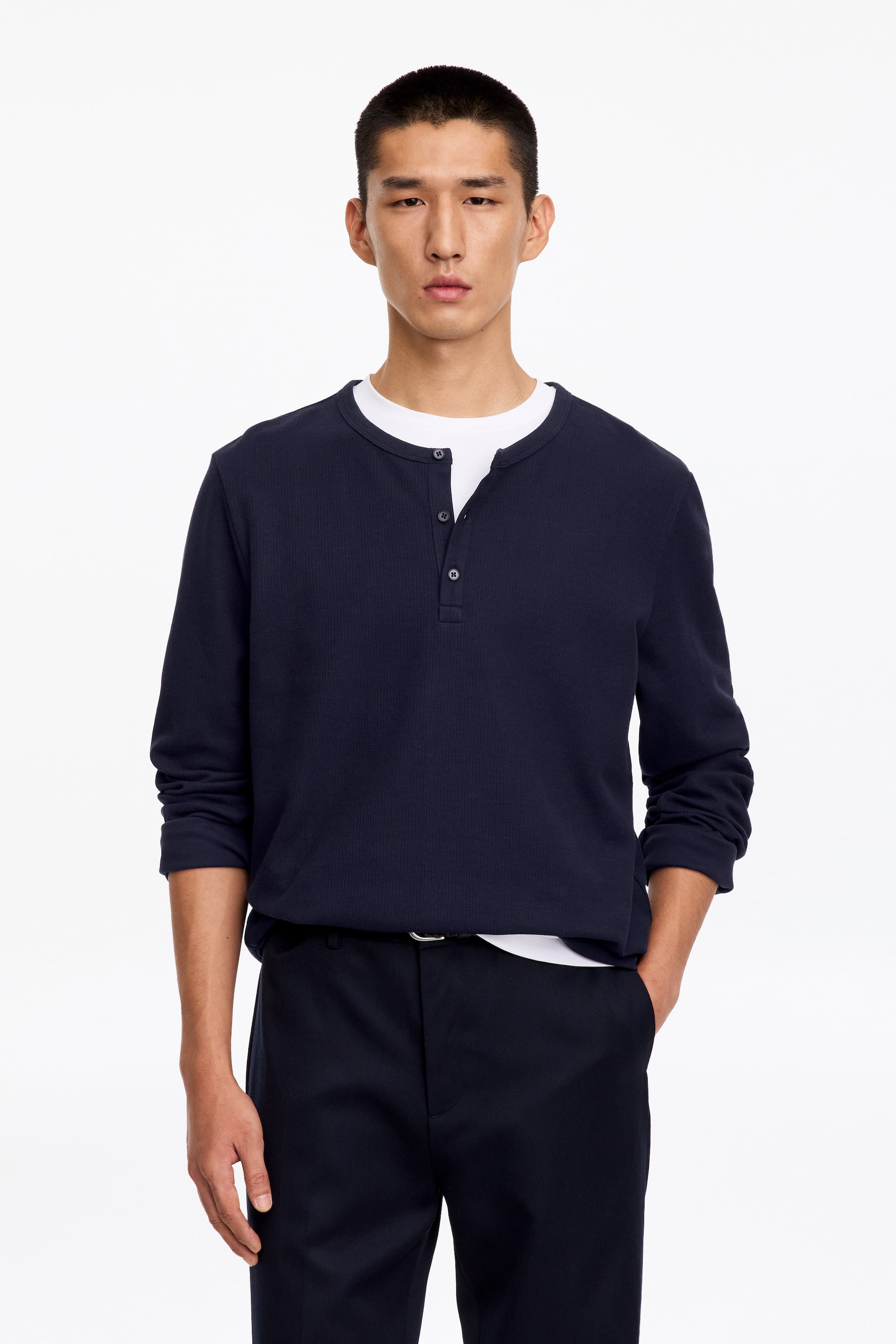 Ver imagen más grande: La modelo lleva una camisa Henley de color azul marino oscuro, confeccionada en un tejido de punto canalé, sobre una camiseta blanca de cuello redondo. La camisa de manga larga presenta un ribete acanalado en el cuello y una tapeta media con botones parcialmente desabrochados, y se combina con un pantalón oscuro.