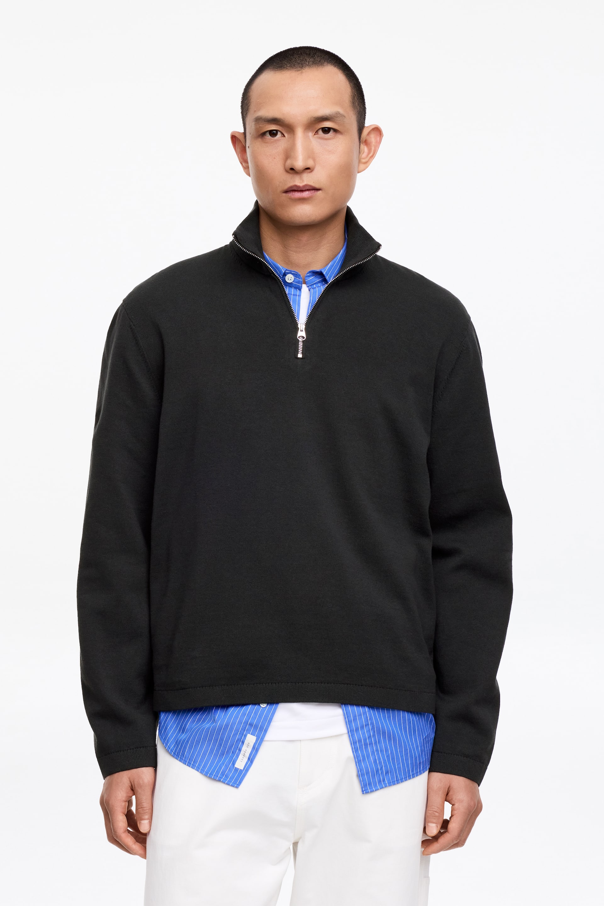 Visa större bild: En svart quarter-zip-tröja presenteras, med stående krage och en silverfärgad dragkedja. Den bärs över en blå- och vitrandig skjorta med krage och en vit t-shirt, vilka syns vid kragen och nederkanten.