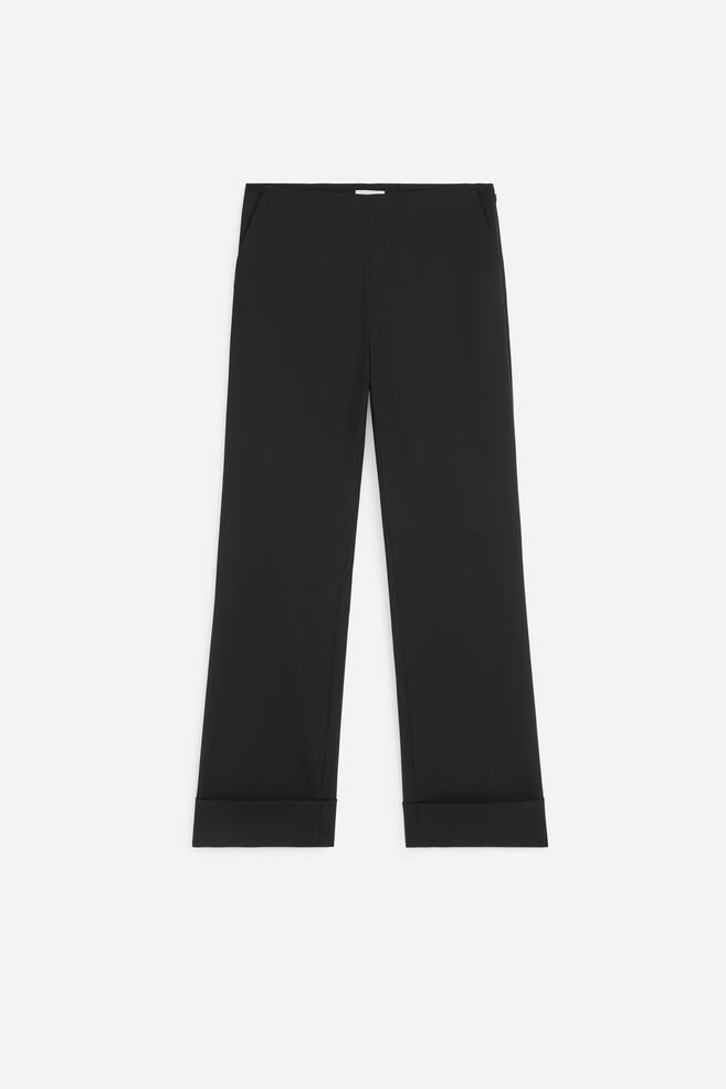 Die schwarze Hose ist in der Frontansicht zu sehen und verfÃ¼gt Ã¼ber einen hohen Bund mit einer klaren, nahtlosen Vorderseite sowie eine gerade Silhouette. Die Hose ist mit breiten, umgeschlagenen SaumaufschlÃ¤gen versehen. - 1