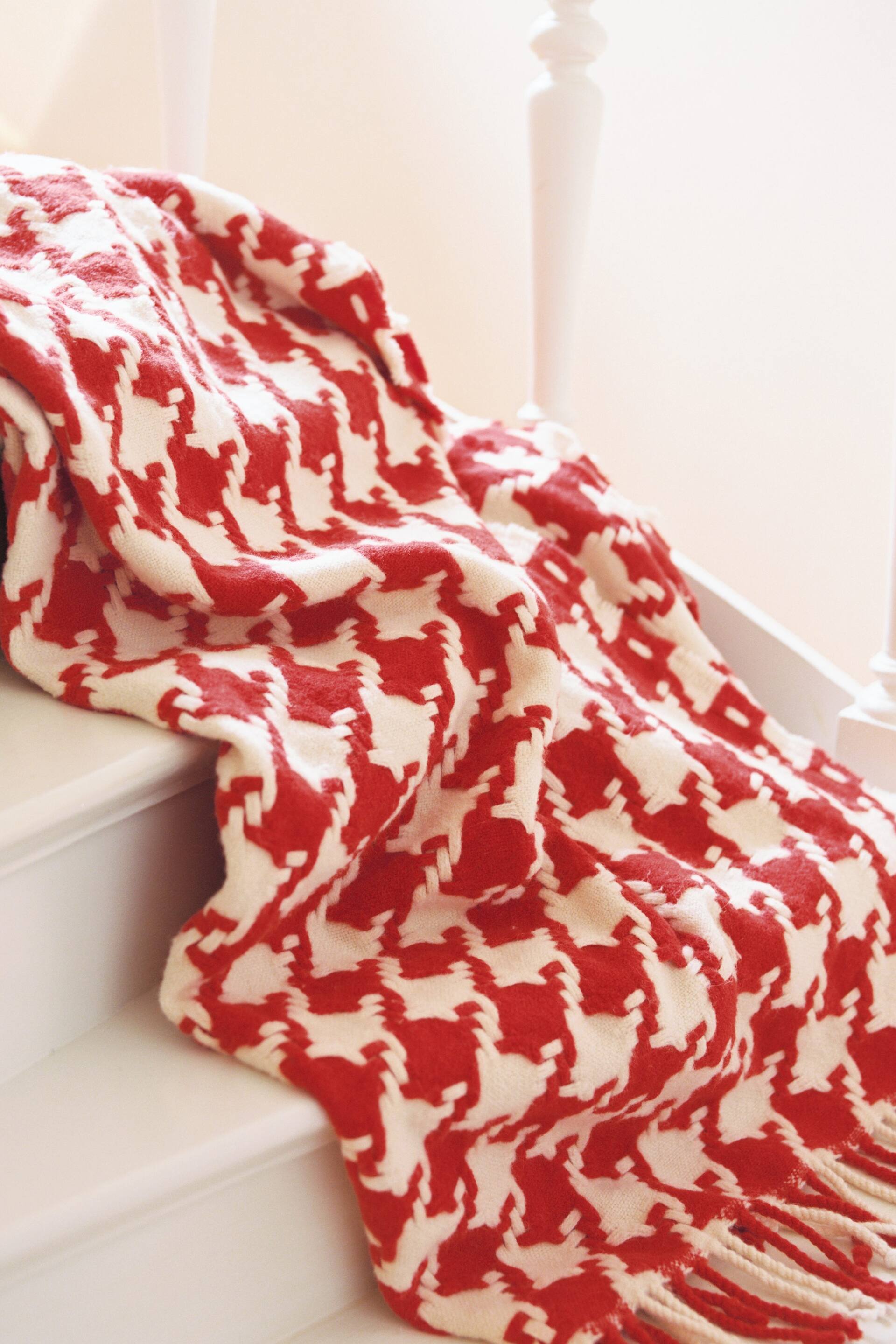 Se større billede: Et rødt og off-white tæppe med et hundetandsmønster, draperet diagonalt og med synlige folder. Frynser pryder dens nederste højre kant.