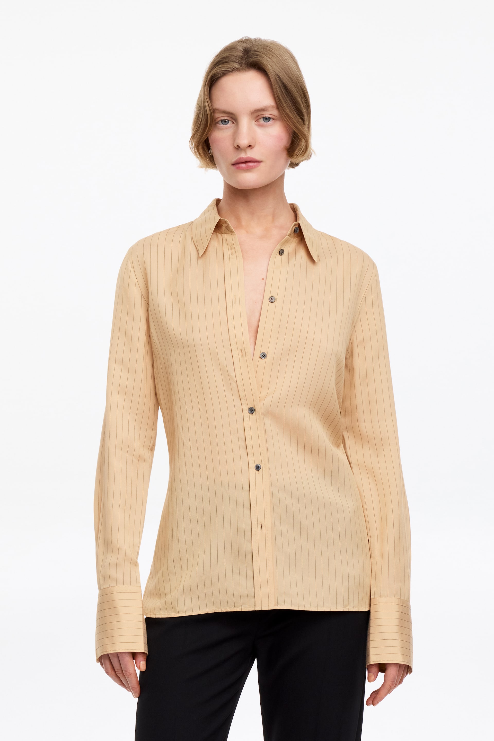 Visualizza l'immagine ingrandita: La camicia a righe beige è presentata di fronte, con un colletto a punta e una lista frontale parzialmente sbottonata. Presenta maniche lunghe con polsini ampi a bottone singolo ed è abbinata a pantaloni neri visibili.