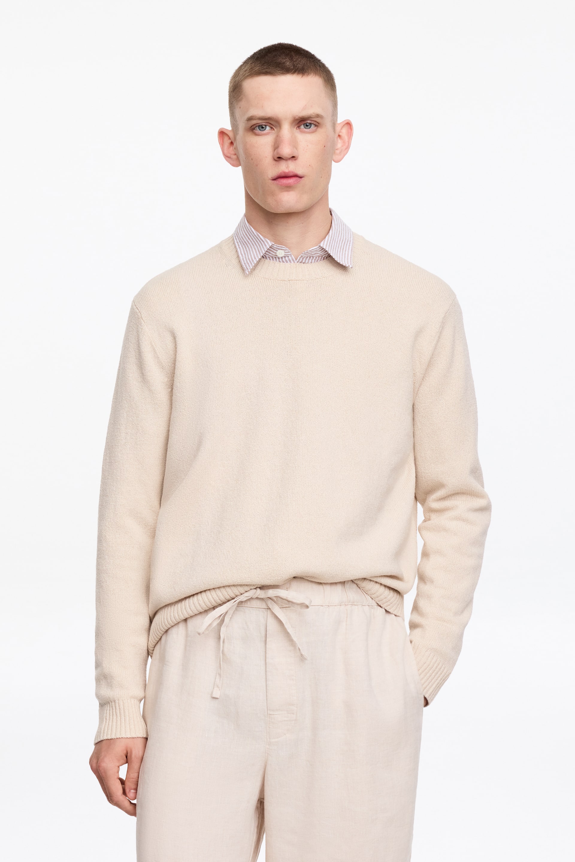 Grösseres Bild ansehen: Ein hellbeiger Pullover mit Rundhalsausschnitt wird über einem gestreiften Hemd mit Kragen getragen, das lange Ärmel mit gerippten Bündchen und einen gerippten Saum aufweist. Hellbeige Leinenhosen verfügen über einen Kordelzug in der Taille und Seitentaschen.