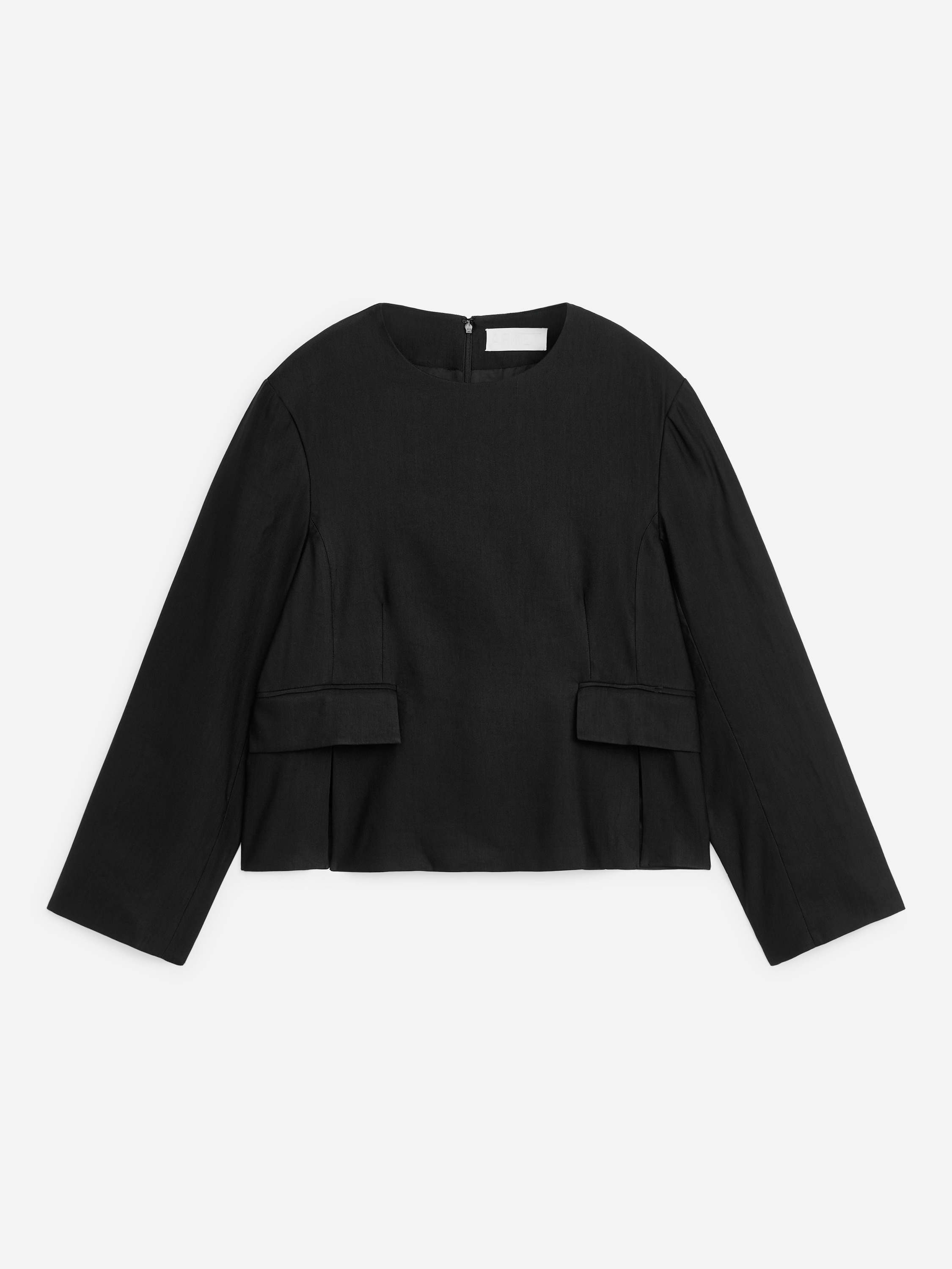 Una camicetta nera strutturata con scollo rotondo, maniche lunghe, patte decorative sulle tasche in vita e spacchi laterali viene indossata dalla modella. La camicetta è abbinata a jeans neri a gamba larga e la modella tiene in mano una piccola borsa nera.