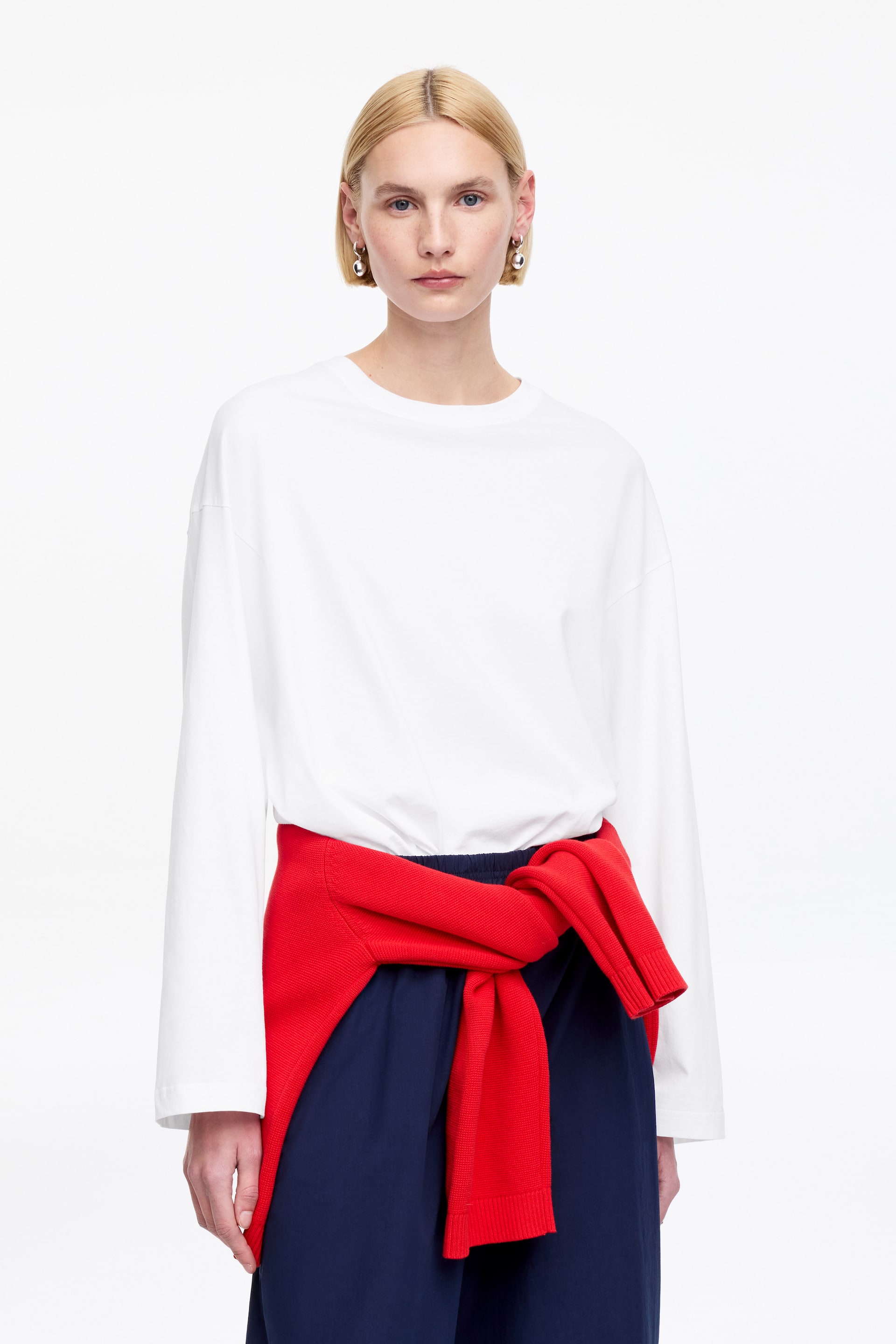 Ingrandisci l'immagine: Un top bianco a maniche lunghe viene presentato, caratterizzato da un girocollo e una vestibilità rilassata e oversize. Le sue maniche lunghe e semplici si estendono fino ai polsi. Un maglione rosso è legato intorno alla vita, oscurando parzialmente la parte inferiore del top.