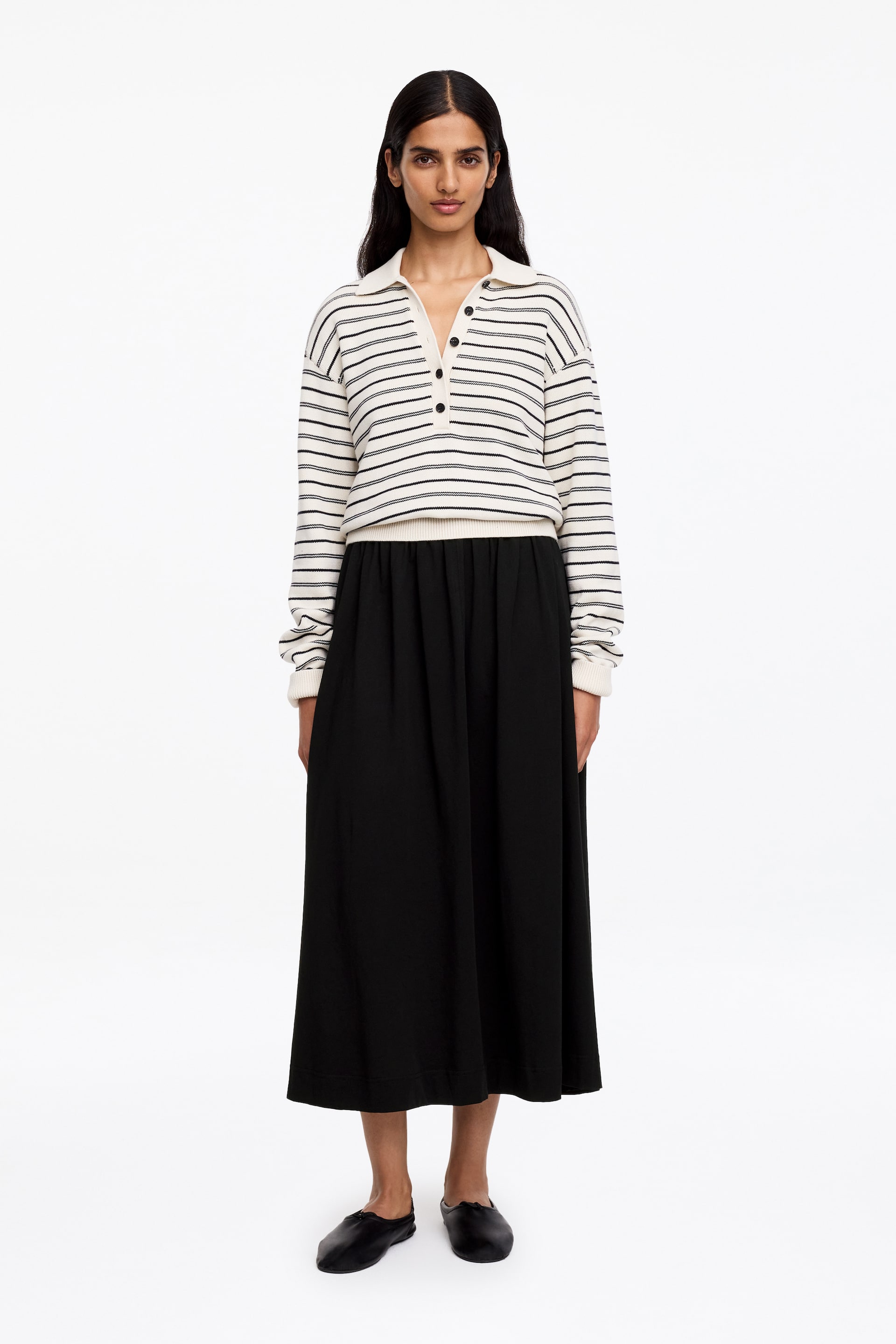 Visualizza immagine più grande: Un maglione polo a maniche lunghe a righe viene presentato, mostrando una base color bianco sporco con righe orizzontali nere, un colletto polo, una chiusura a tre bottoni e polsini e orlo a coste. Viene indossato con una gonna midi nera che cade in una silhouette fluida a trapezio da una vita arricciata.
