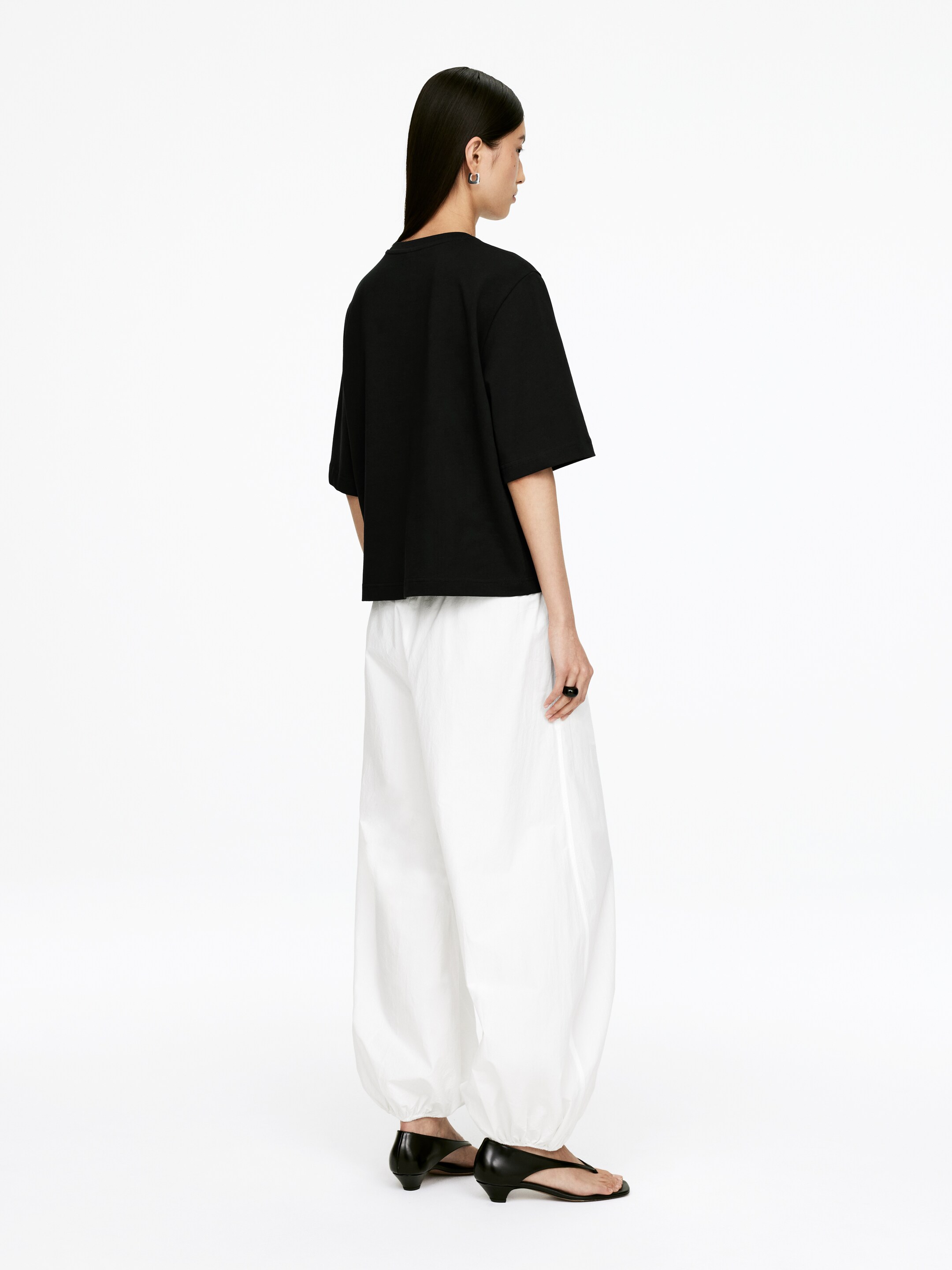 View larger image: A black, relaxed-fit t-shirt is being presented from a back-three-quarter angle, featuring short, wide sleeves and a straight, cropped hem. It is being styled with white, voluminous pants that are ballooning through the leg and gathering at the ankles.