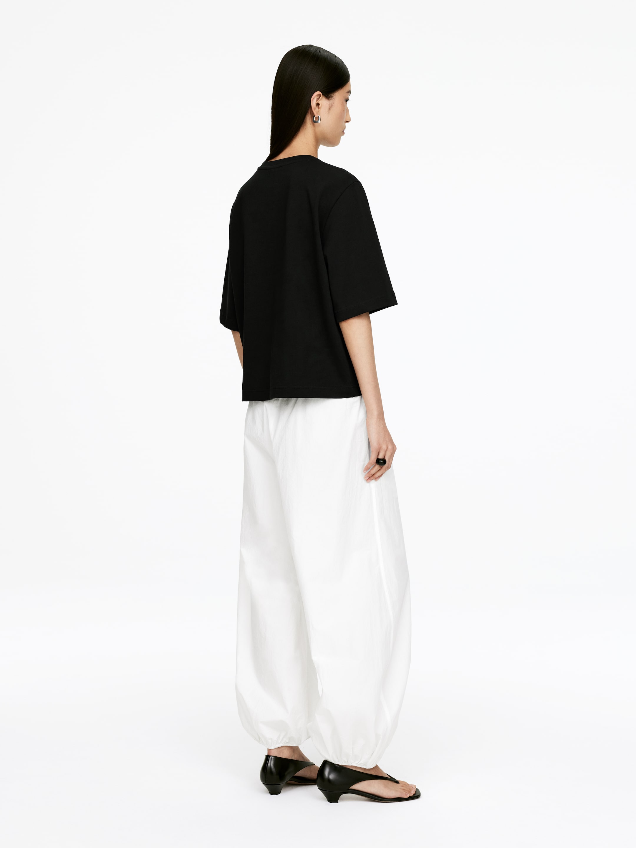 Grotere afbeelding bekijken: Een zwart, relaxed-fit t-shirt wordt gepresenteerd vanuit een driekwart achteraanzicht, met korte, wijde mouwen en een rechte, cropped zoom. Het wordt gestyled met witte, volumineuze broeken die door het been balloneren en bij de enkels samenkomen.