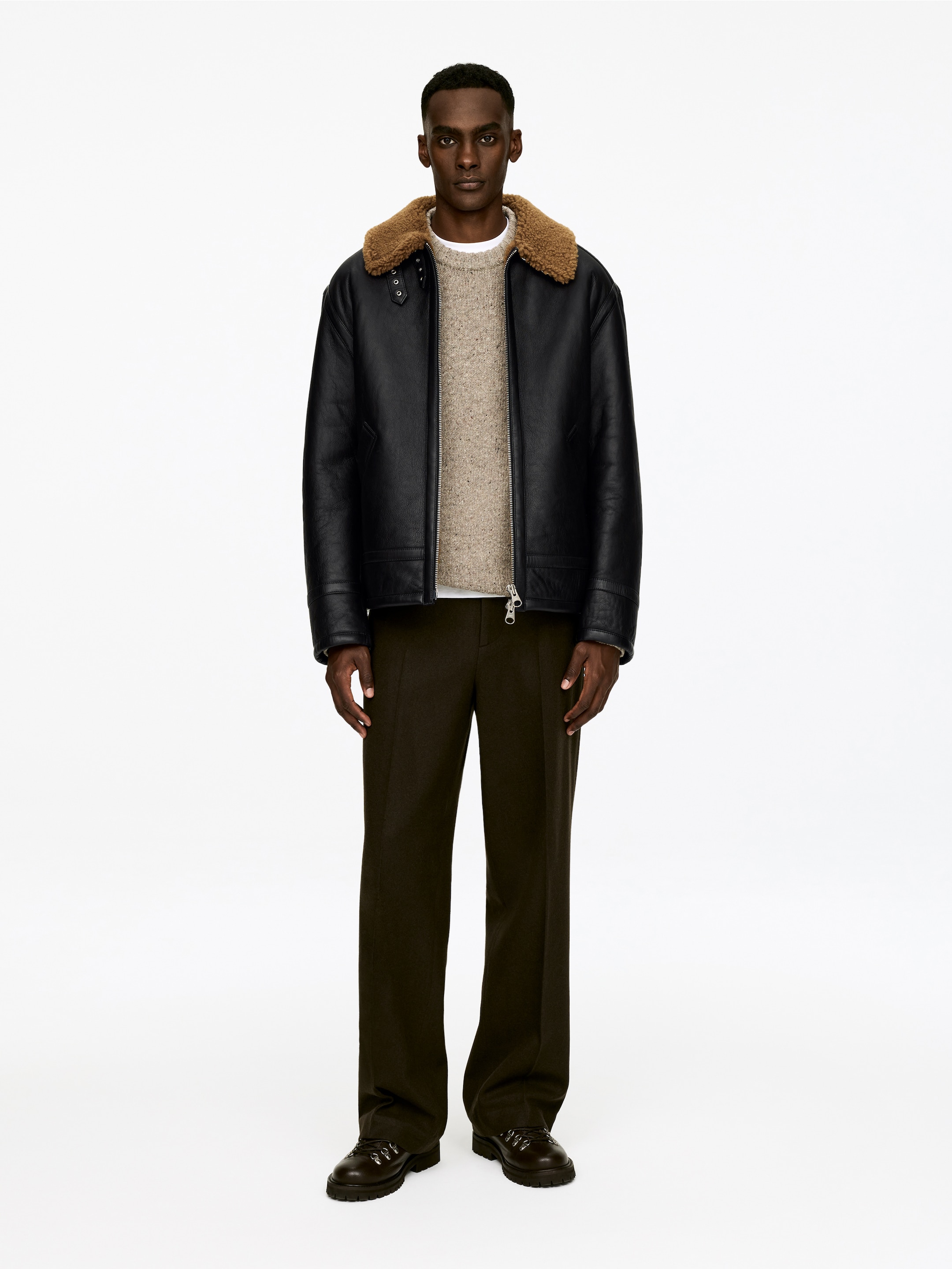 Agrandir l'image: Une veste en cuir noir est présentée de face. Elle est dotée d'un col en shearling marron avec deux sangles à boucle, d'une fermeture éclair sur le devant et de poches latérales zippées. La veste est portée par-dessus un pull texturé de couleur claire.