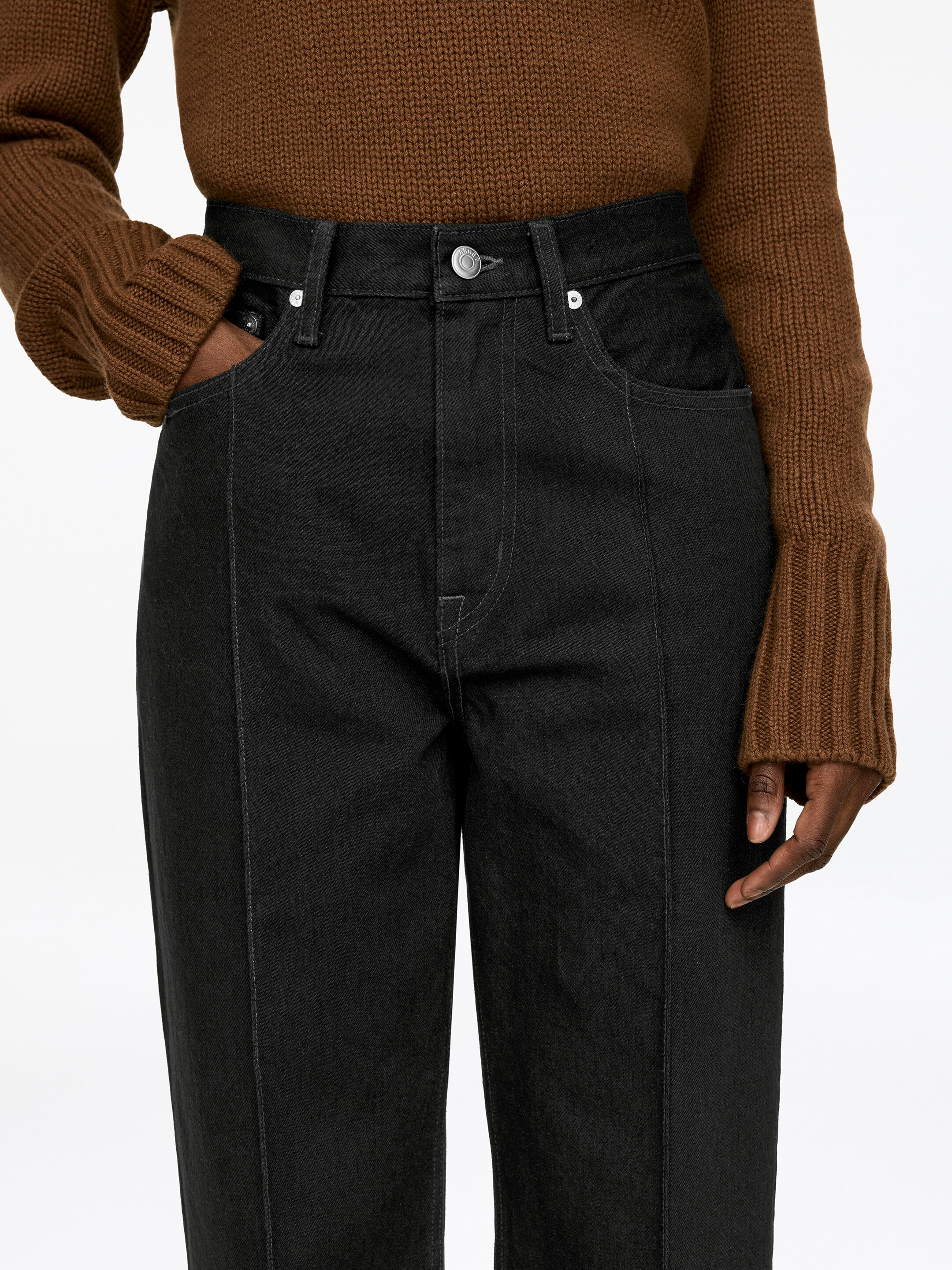 Agrandir l'image: Un jean noir est présenté de face, légèrement en biais, avec une taille haute, un bouton visible et des passants de ceinture. Le jean montre des poches avant avec des rivets et des coutures verticales distinctes qui descendent le long de chaque jambe. Des coutures contrastées blanches mettent en valeur ces coutures et d'autres détails de construction.