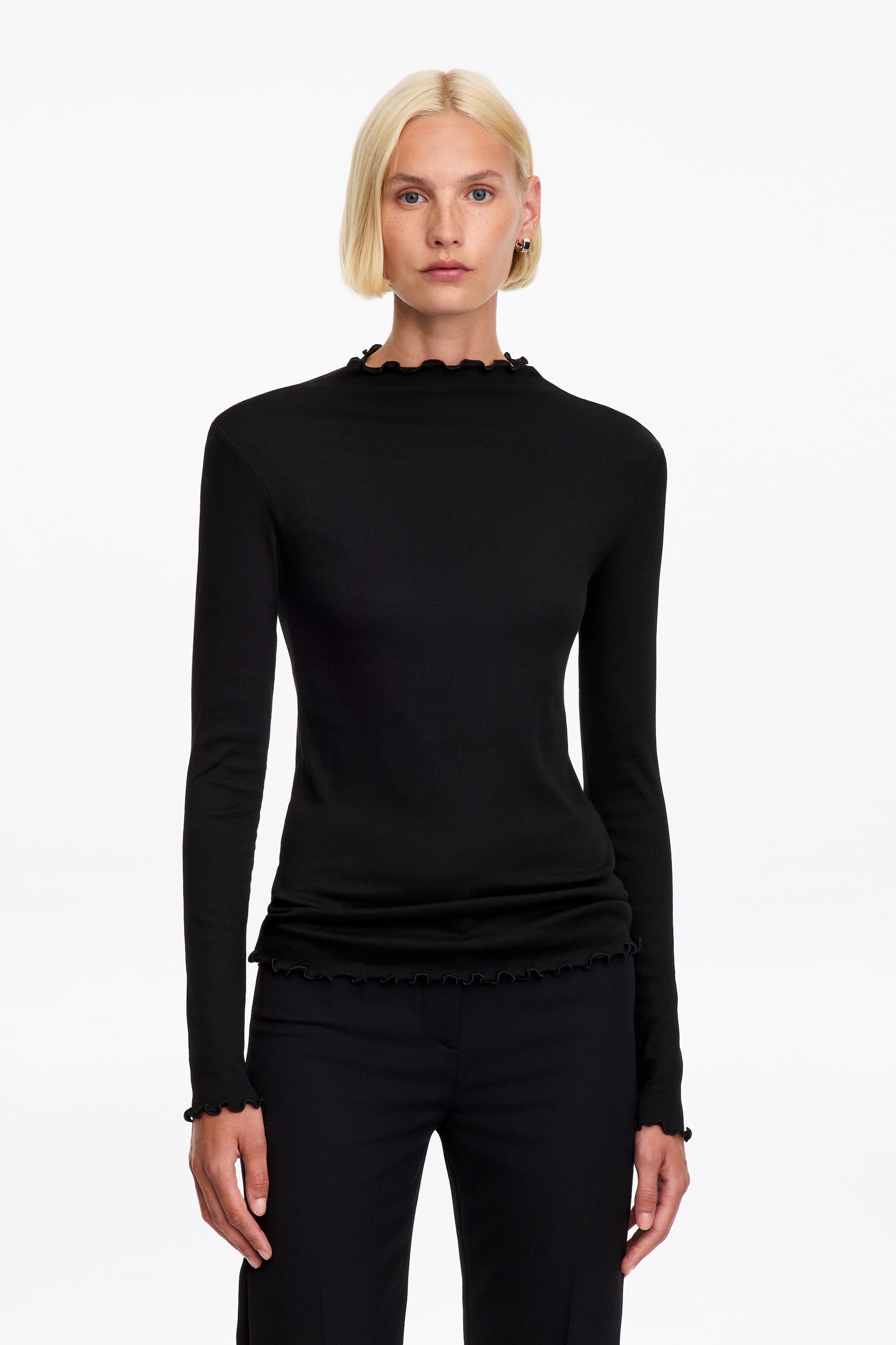 Se større billede: En sort, slim-fit, langærmet jerseytop bæres af modellen, stylet med sorte bukser. Toppen har en høj halsudskæring og fine flæsedetaljer ved kraven, manchetterne og kanten.