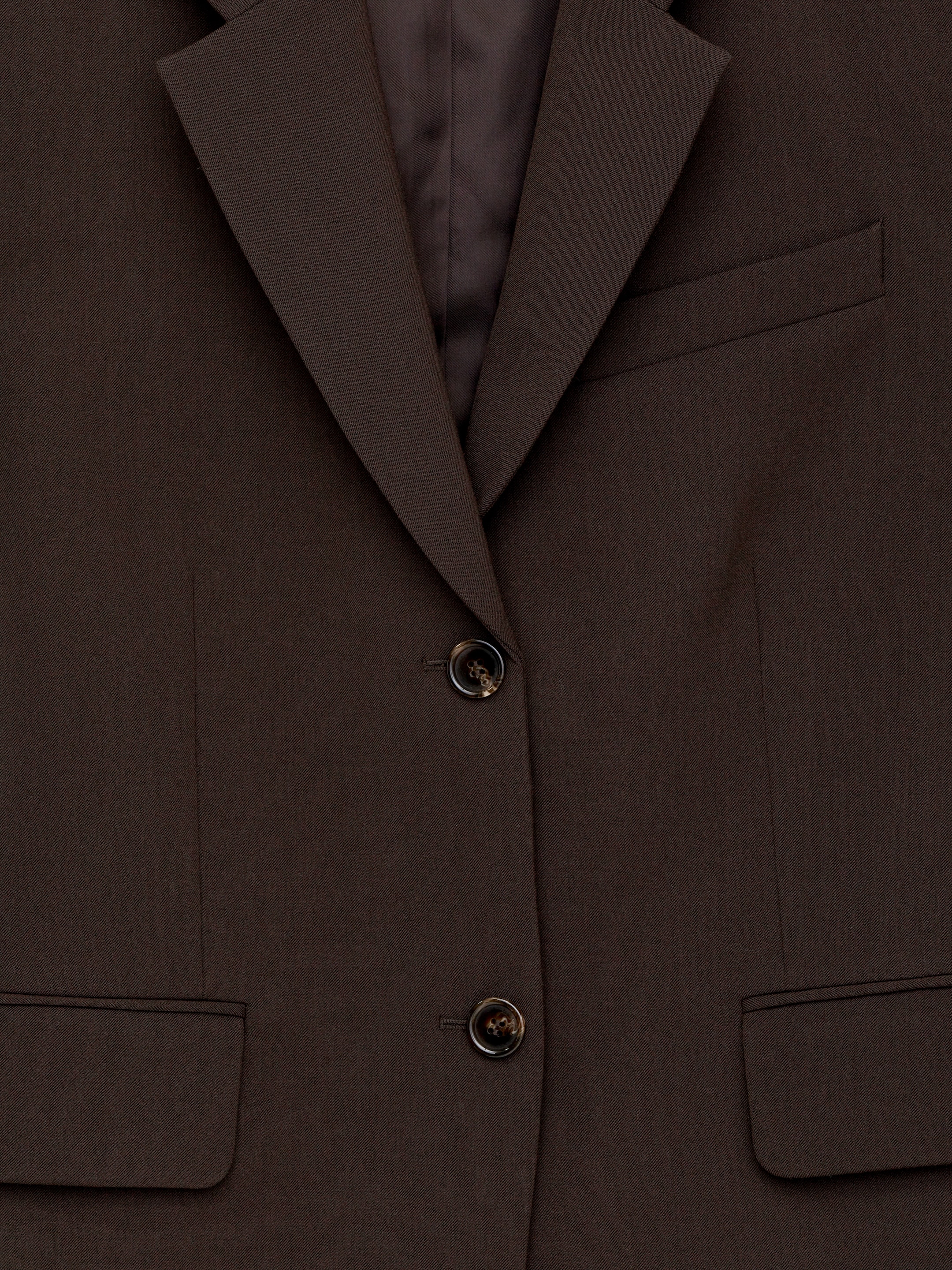 Größeres Bild anzeigen: Ein dunkelbrauner einreihiger Blazer zeigt seine Vorderseite, mit gekerbten Revers und einem sichtbaren dunkler braunen Futter. Er wird durch zwei gesprenkelte dunkelbraune Knöpfe gesichert und verfügt über eine einzelne Leistentasche auf der oberen Brust und zwei Pattentaschen an der unteren Vorderseite.