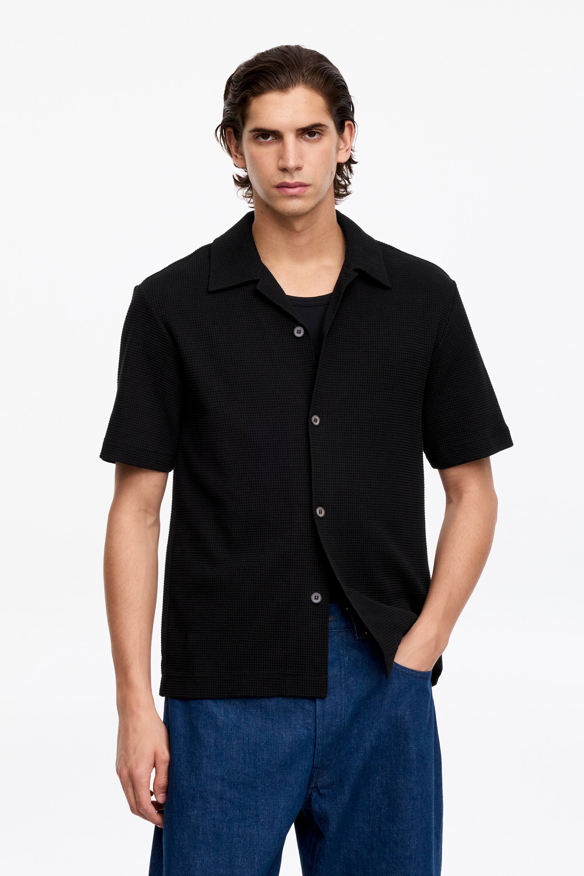 Преглед на по-голямо изображение: Черна риза с къс ръкав и текстурирана плетка тип "вафла", представена с разтворена яка и предно закопчаване със светлосиви копчета, носена разкопчана върху черна тениска.