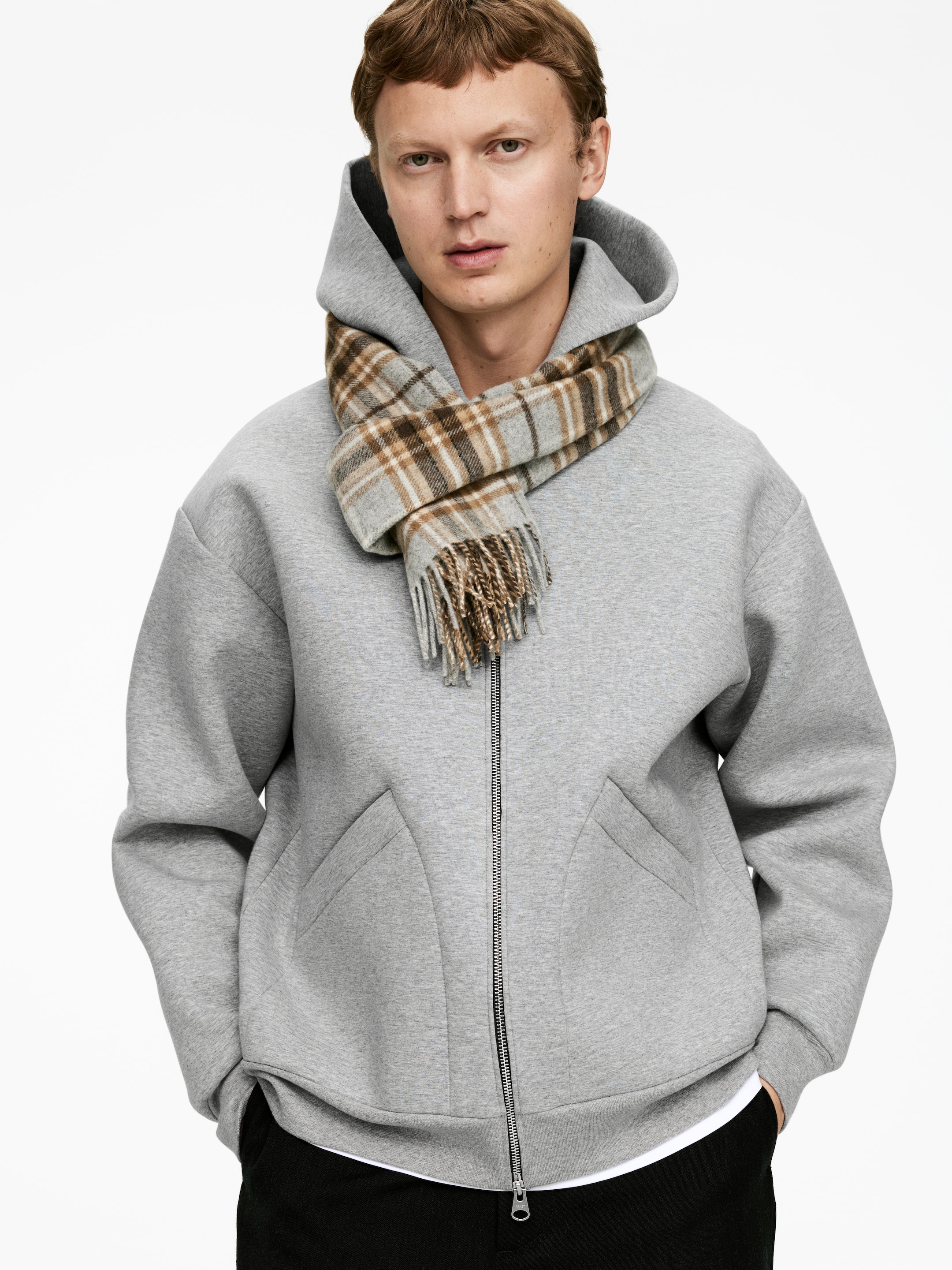 View larger image: A light grey structured hoodie is being presented front-facing, featuring a full-length front zipper and two distinct, angular front pockets. A grey and brown plaid scarf with fringed ends is being layered over the hoodie, wrapped around the neck.