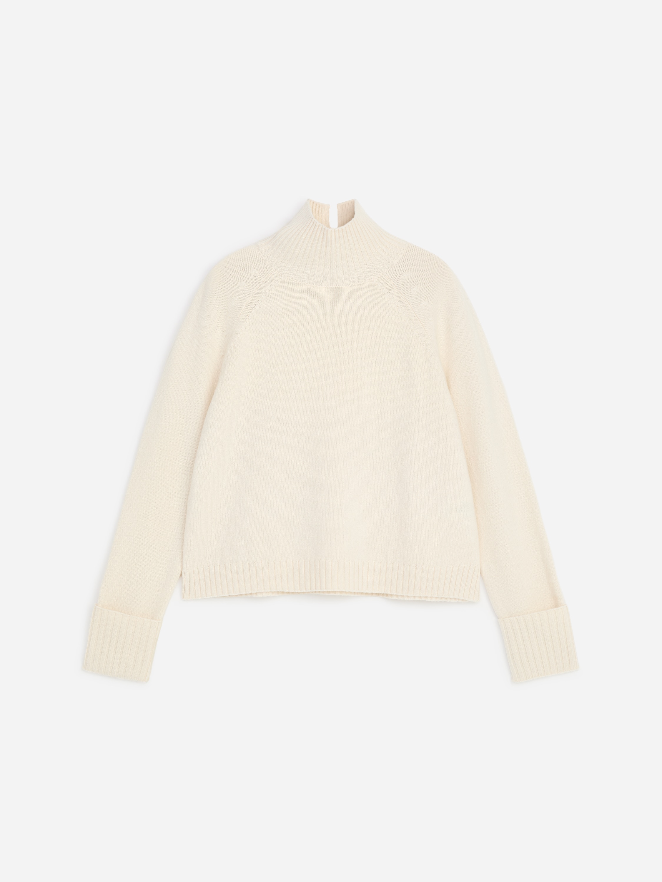 La modella indossa un maglione lavorato a maglia color bianco sporco con collo alto risvoltato, caratterizzato da maniche raglan, polsini a coste risvoltati e fondo a coste. Il maglione è abbinato a pantaloni bianchi, con la modella che tiene una mano in tasca.