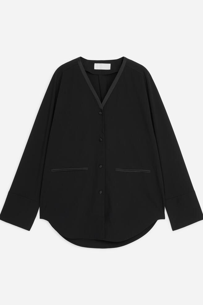 Ein schwarzer Tuxedo-Blazer im entspannten Schnitt ist von vorne zu sehen und zeigt einen kragenlosen V-Ausschnitt mit kontrastierendem Satinbesatz sowie einen Knopfverschluss mit stoffbezogenen KnÃ¶pfen. Das Design verfÃ¼gt Ã¼ber lange Ãrmel mit breiten Manschetten, zwei doppelte Leistentaschen mit Satinbesatz und einen geschwungenen Saum. - 1