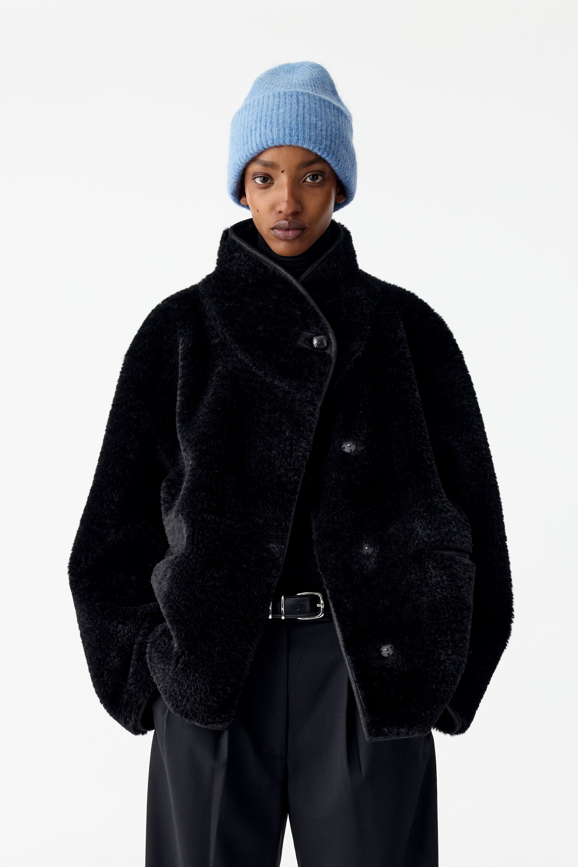 Größeres Bild ansehen: Eine schwarze, strukturierte Shearling-Jacke mit hohem Kragen und asymmetrischer Knopfleiste wird vom Model getragen, das mit den Händen in den Seitentaschen steht. Die Jacke ist mit einem schwarzen Rollkragenpullover, schwarzen Hosen mit einem Gürtel mit silberner Schnalle und einer hellblauen Rippstrick-Beanie kombiniert.