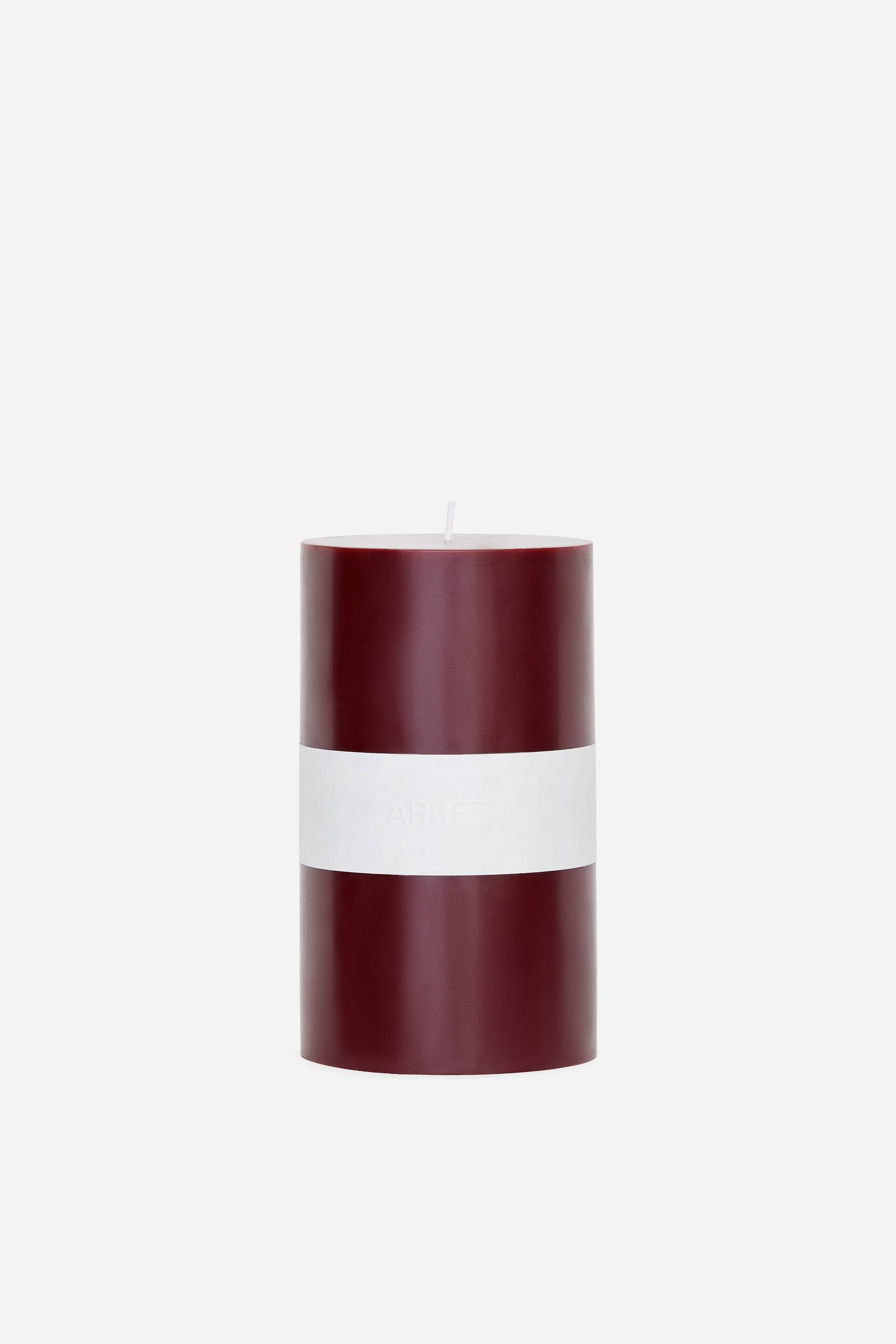 View larger image: A dark red wax pillar candle is standing facing forward, featuring a cylindrical shape with a single white wick protruding from its flat top. A wide white paper band wraps around the middle section of the smooth wax candle.