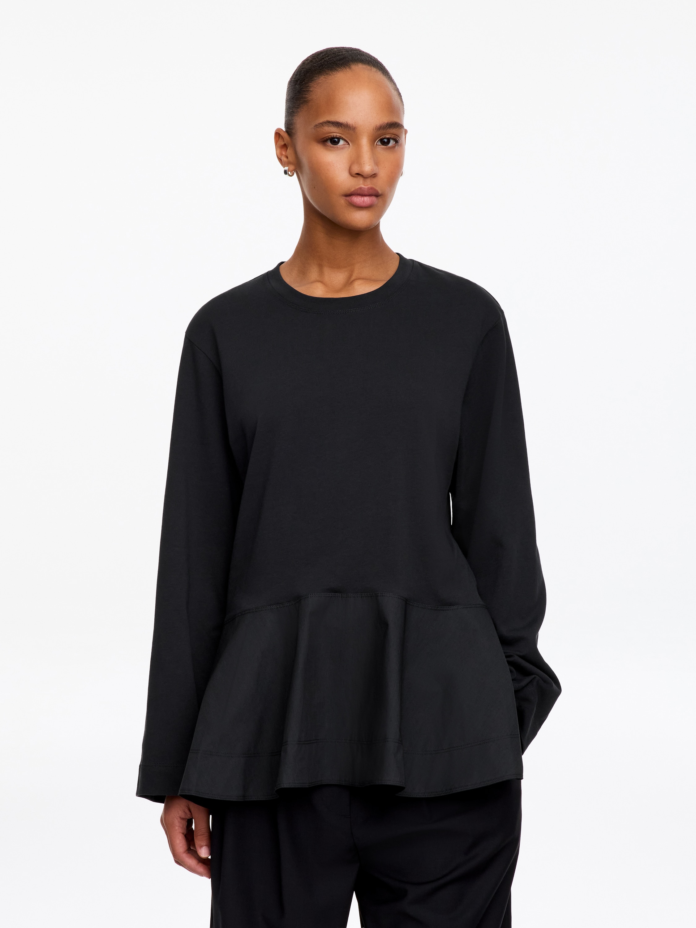 A black long-sleeved top is shown from the front, featuring a matte jersey upper body with a classic crew neckline and wide, straight sleeves. The design transitions into a gathered, flared peplum hem made from a contrasting, slightly textured woven fabric.