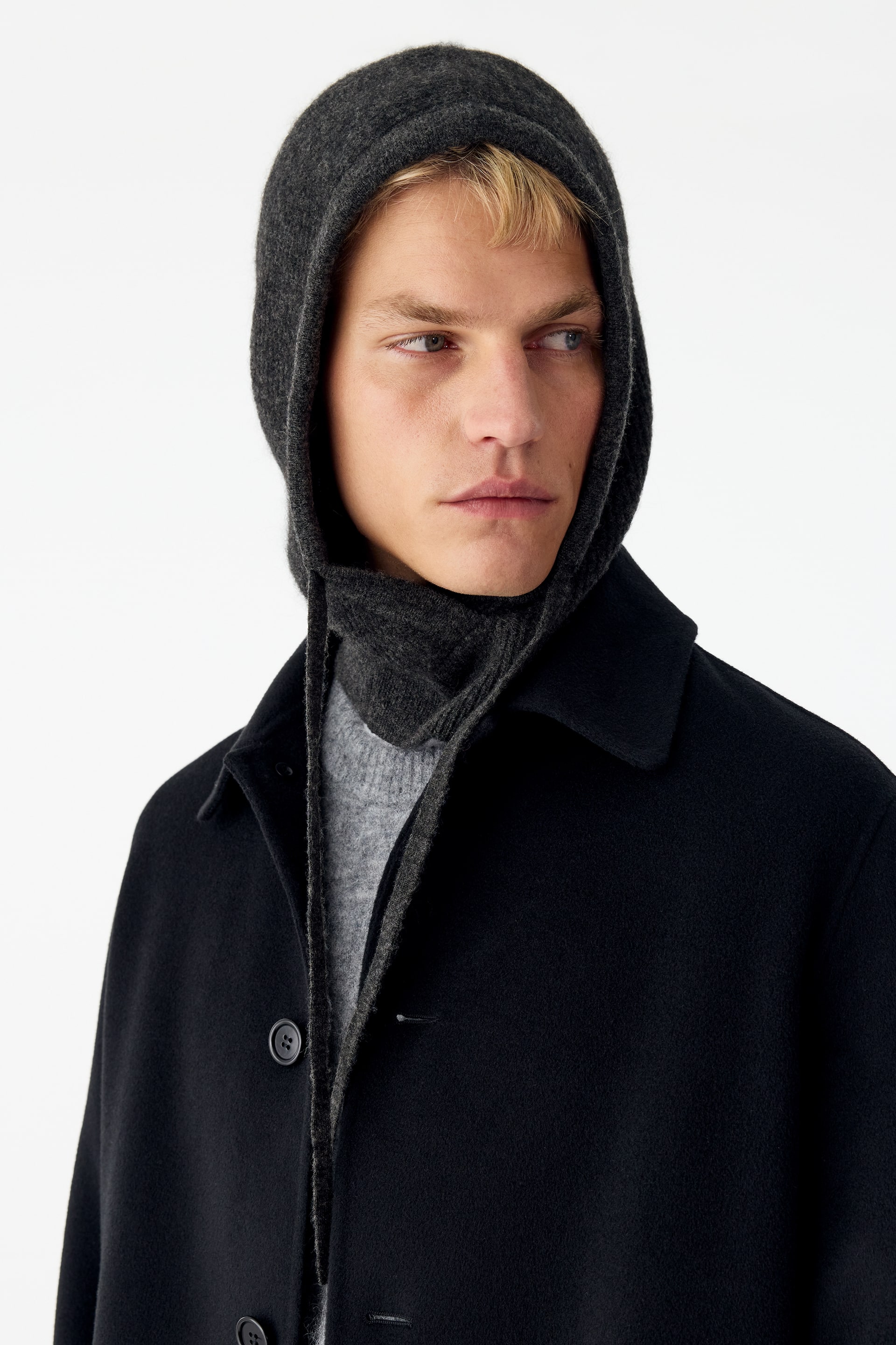 Grotere afbeelding bekijken: Het model draagt een donkergrijze gebreide capuchon die het hoofd en de nek bedekt, met lange bijpassende trekkoorden die aan de voorkant naar beneden hangen. De capuchon is gelaagd gestyled over een lichtgrijze sweater met ronde hals en onder een zwarte jas met kraag.