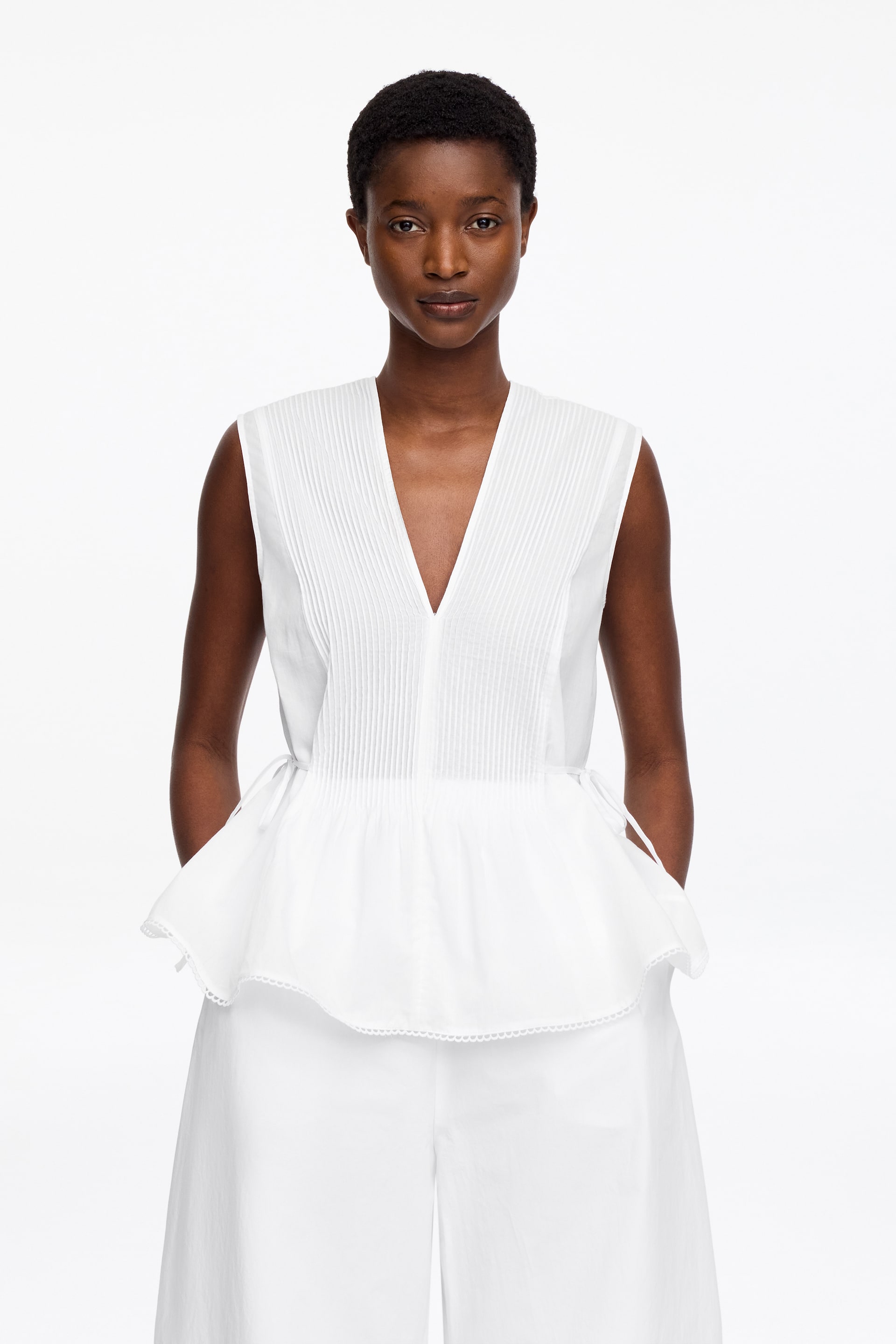 Se større billede: En hvid ærmeløs top med en dyb V-udskæring og en plisseret overdel, der snøres i taljen med sidestropper og breder sig ud i en peplumkant med bølget kant.