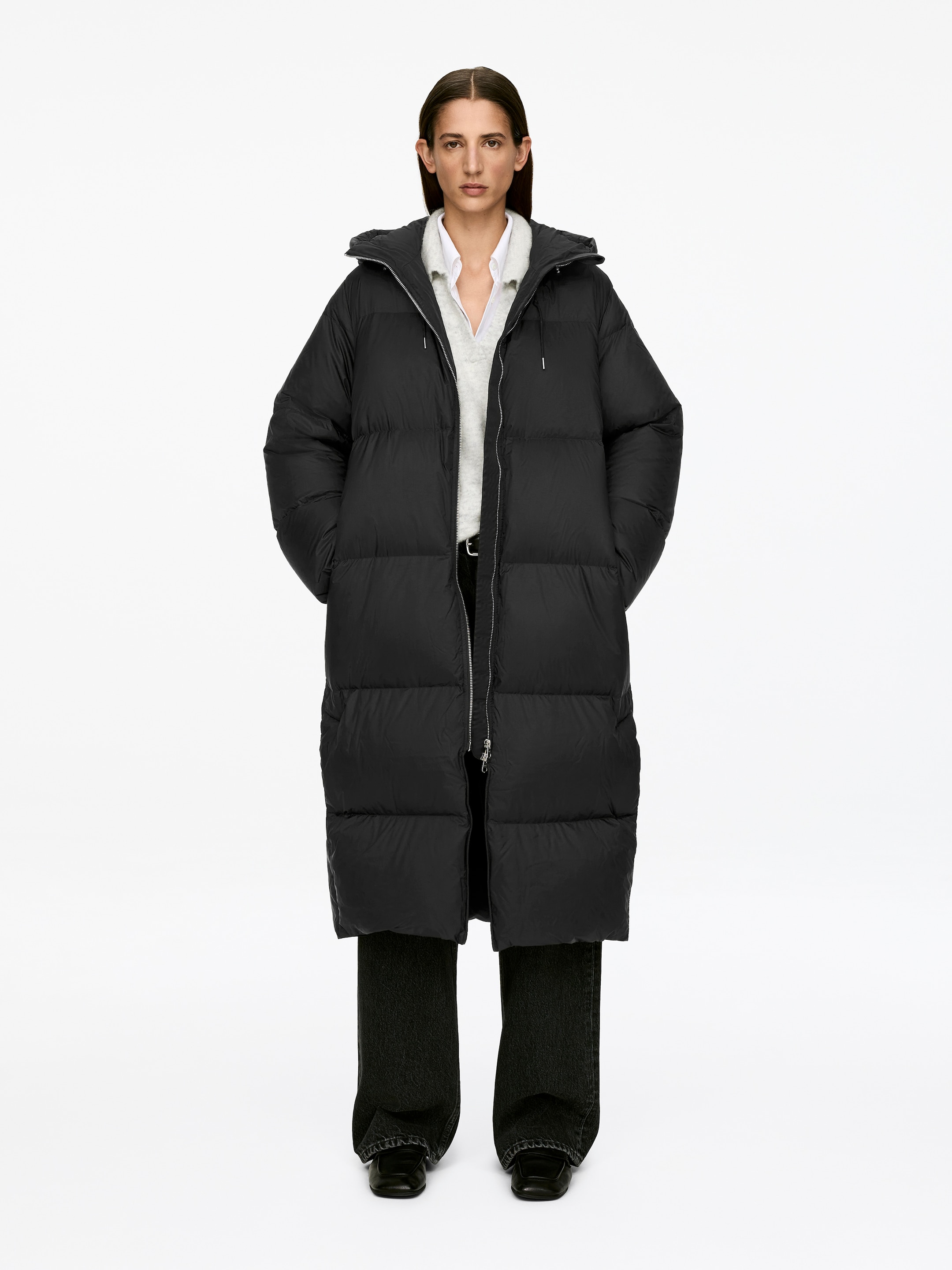 View larger image: Upcycled Down Long Coat in black. The model is wearing the ankle-length puffer coat, which features horizontal quilting and a hood. Its front zipper is partially open, revealing a white shirt and gray sweater, and the model's hands are in its side pockets.