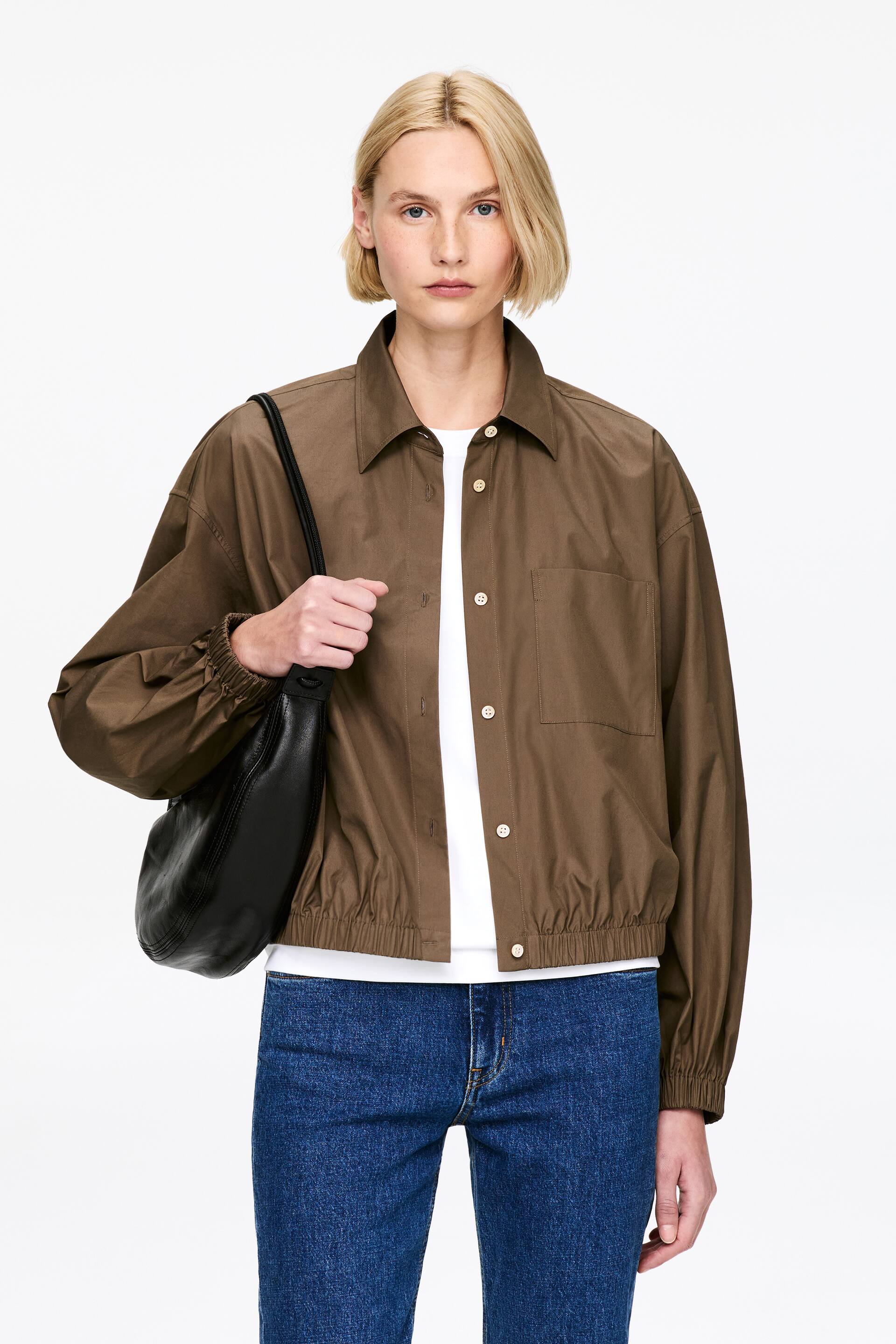 Ver imagen más grande: Una chaqueta marrón que presenta un cuello en punta y un cierre frontal completo con botones de color claro. Está diseñada con mangas largas, puños fruncidos, un dobladillo elástico y un solo bolsillo de parche en el pecho izquierdo, presentado de frente.