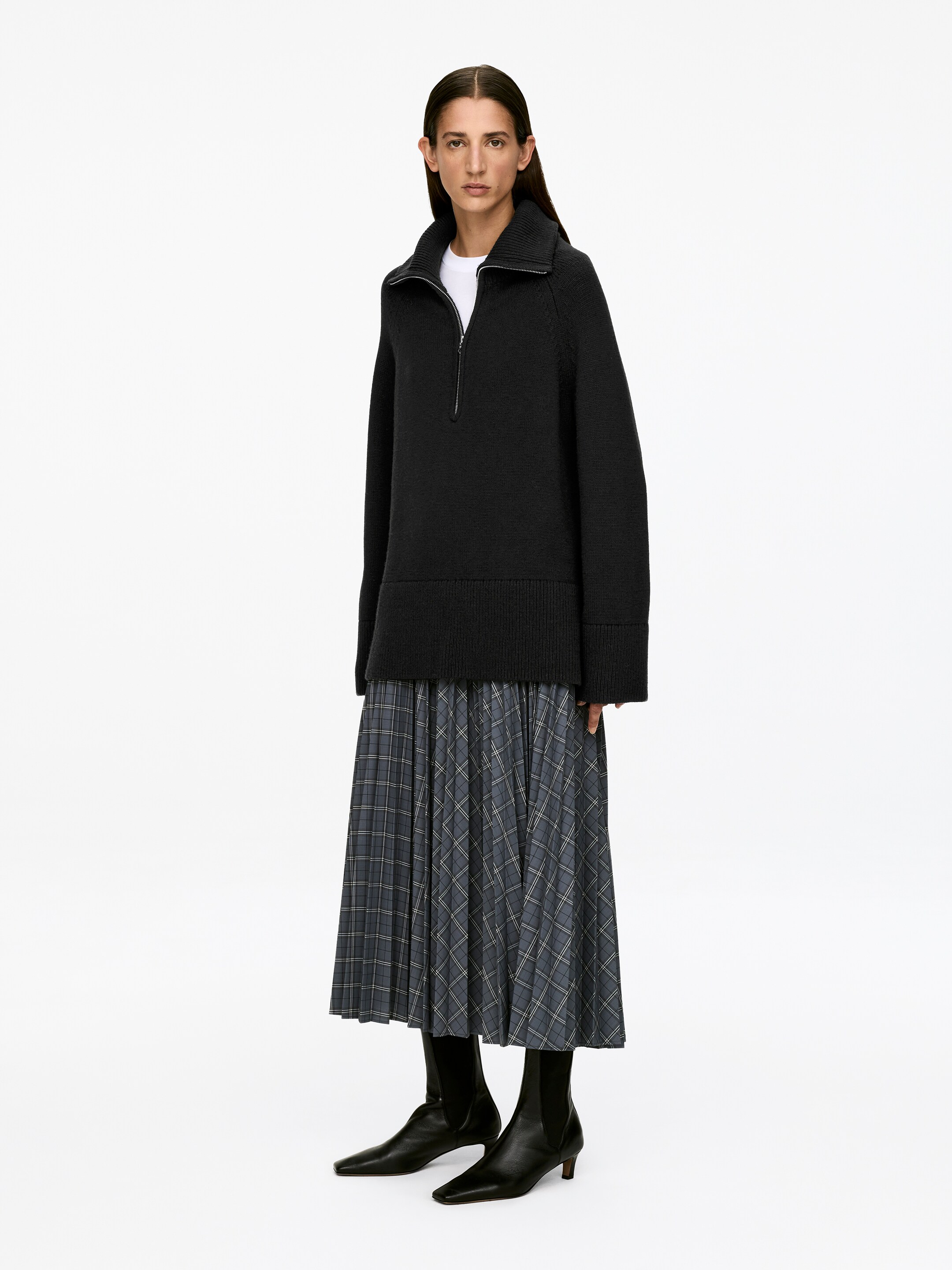 Visualizza immagine più grande: Un maglione nero con mezza zip, caratterizzato da una vestibilità comoda e una cerniera color argento sul colletto alto, è indossato dalla modella. È stratificato su una t-shirt bianca.