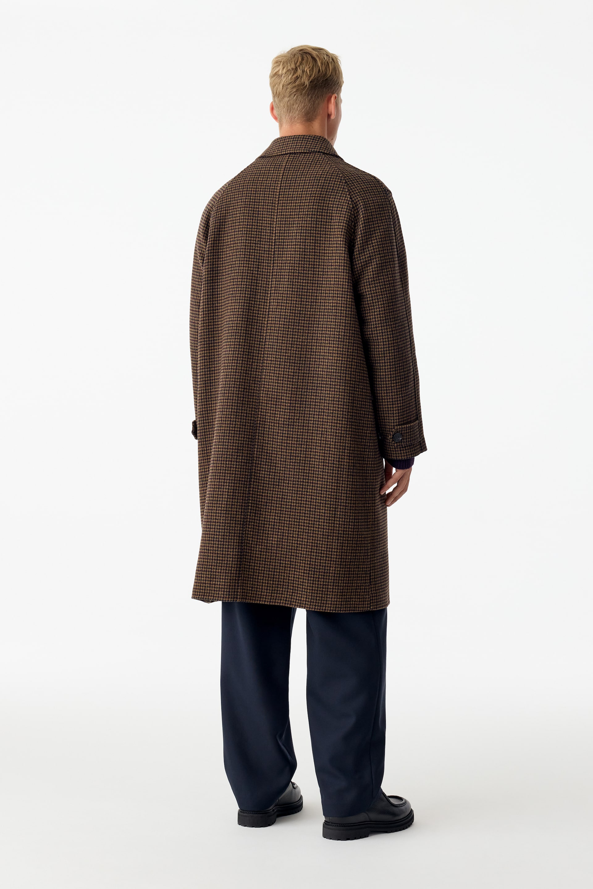 Visualizza immagine più grande: Un cappotto in misto lana a quadri marroni e neri è indossato dalla modella, che è in piedi di spalle alla fotocamera. Il cappotto al ginocchio presenta un colletto classico e dettagli ai polsini con bottoni, ed è abbinato a pantaloni blu scuro e scarpe nere.