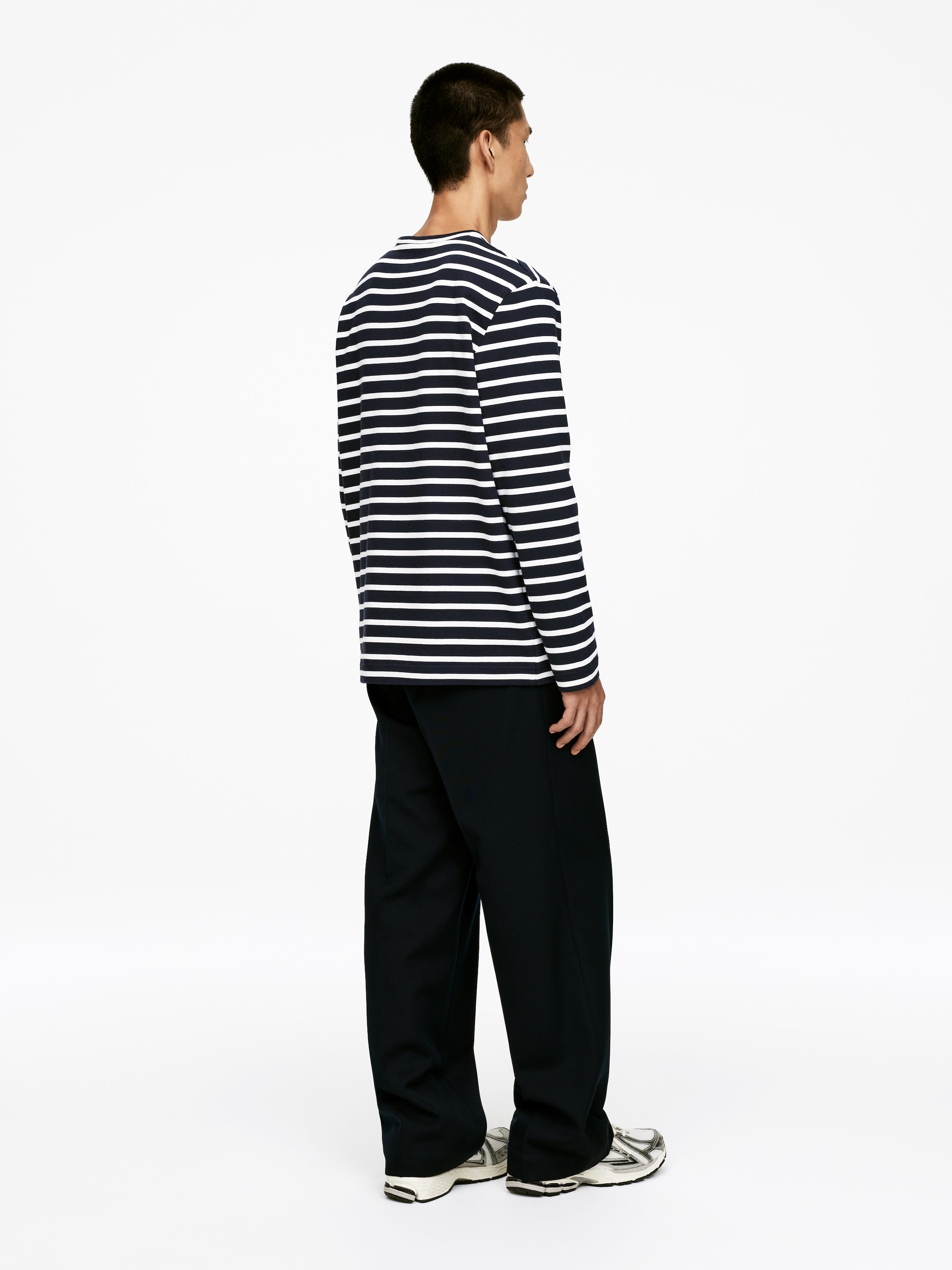 Grösseres Bild ansehen: Ein langärmeliges Rundhals-Shirt mit horizontalen marineblauen und weißen Streifen wird getragen. Das Shirt zeigt eine entspannte Passform und wird mit dunklen, weit geschnittenen Hosen gestylt, aus einer rechten Profilansicht betrachtet.