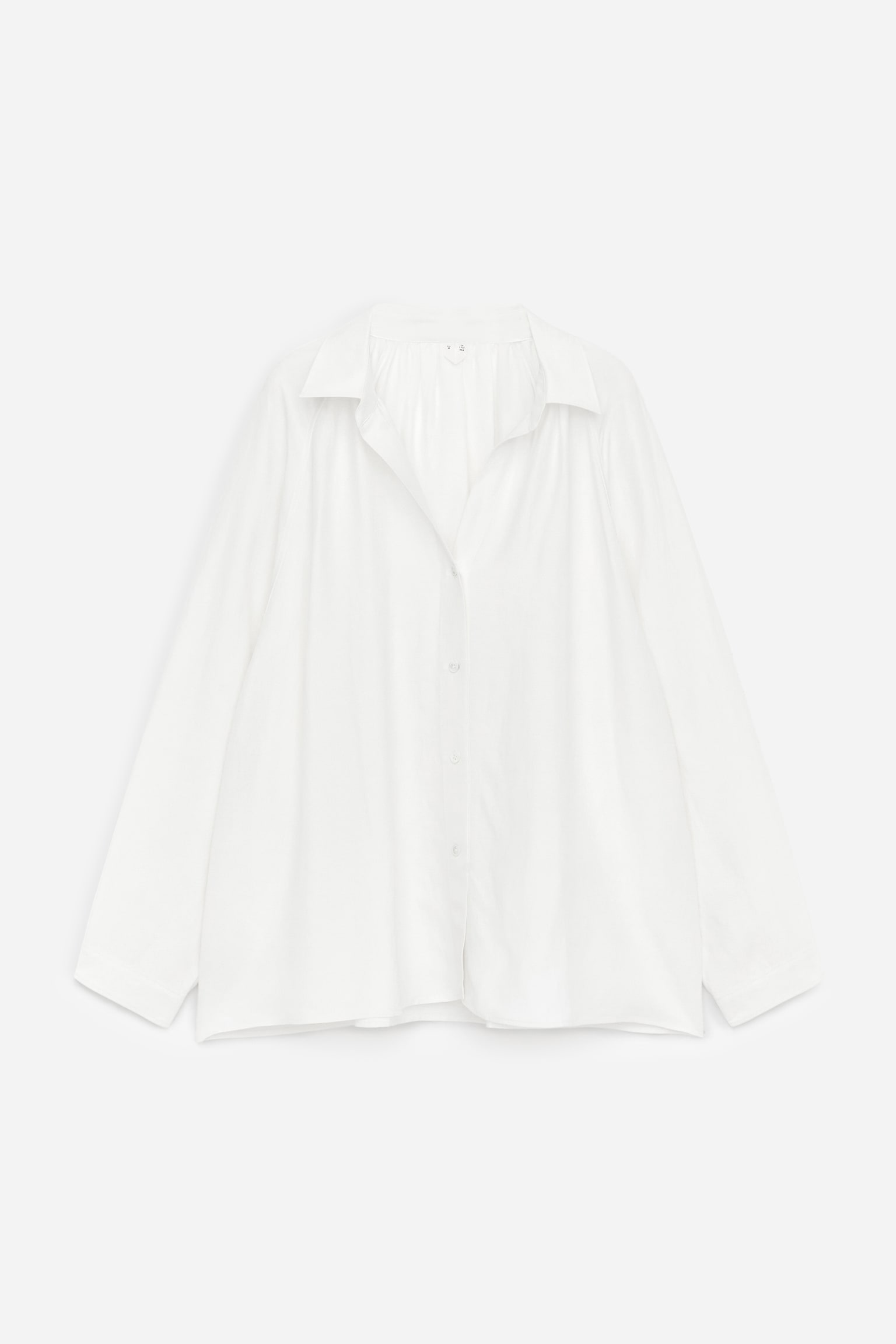 En hvid skjorte præsenterer sin front, med et voluminøst snit og rynker ved halsudskæringen. Stoffet falder blødt og viser en afslappet krave, en fuld knaplukning med hvide knapper og lange ærmer.