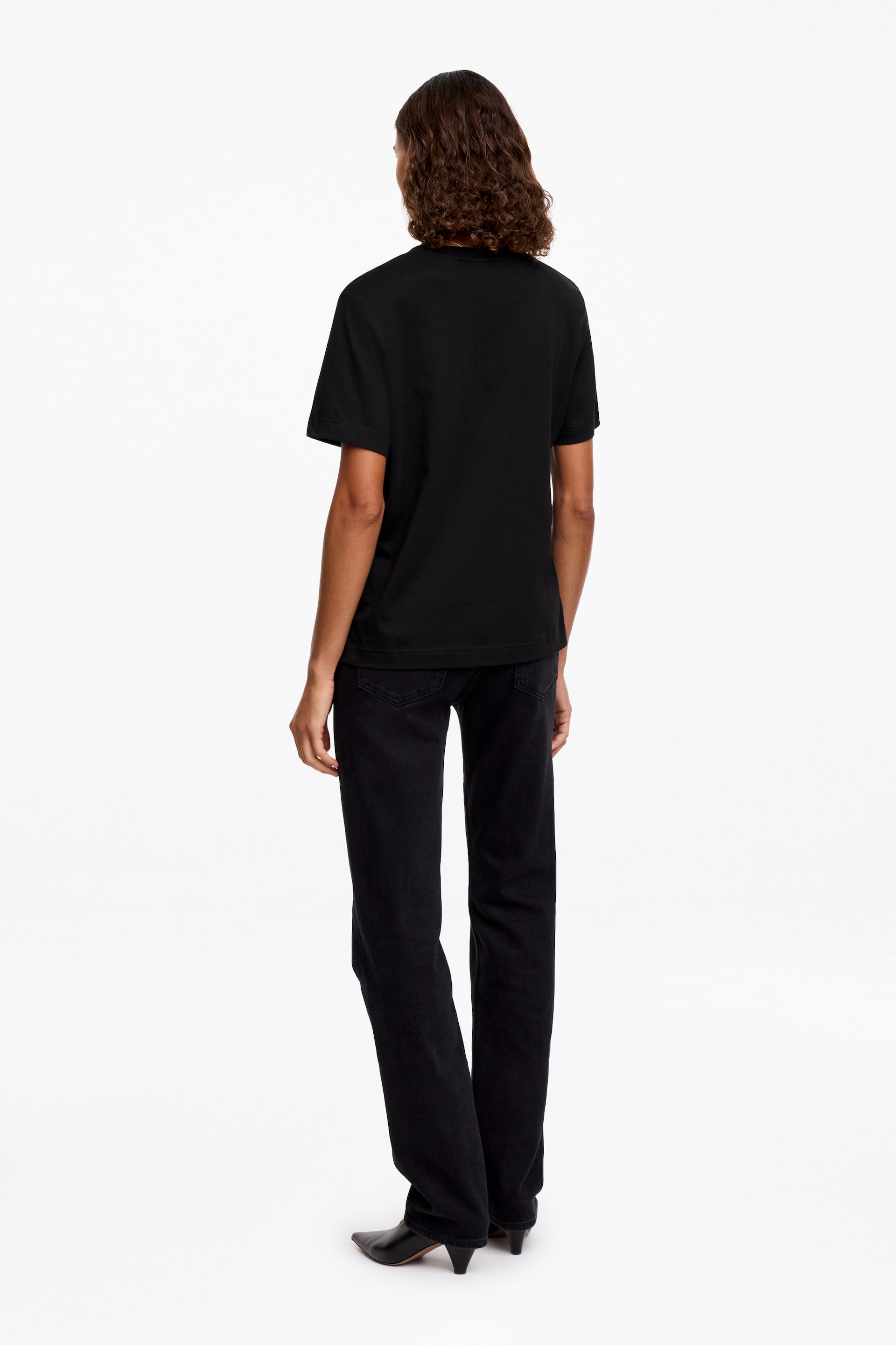 Ver imagem maior: Uma t-shirt preta de manga curta com decote redondo e bainha reta é mostrada de costas, usada com jeans pretos de perna reta com bolsos traseiros.