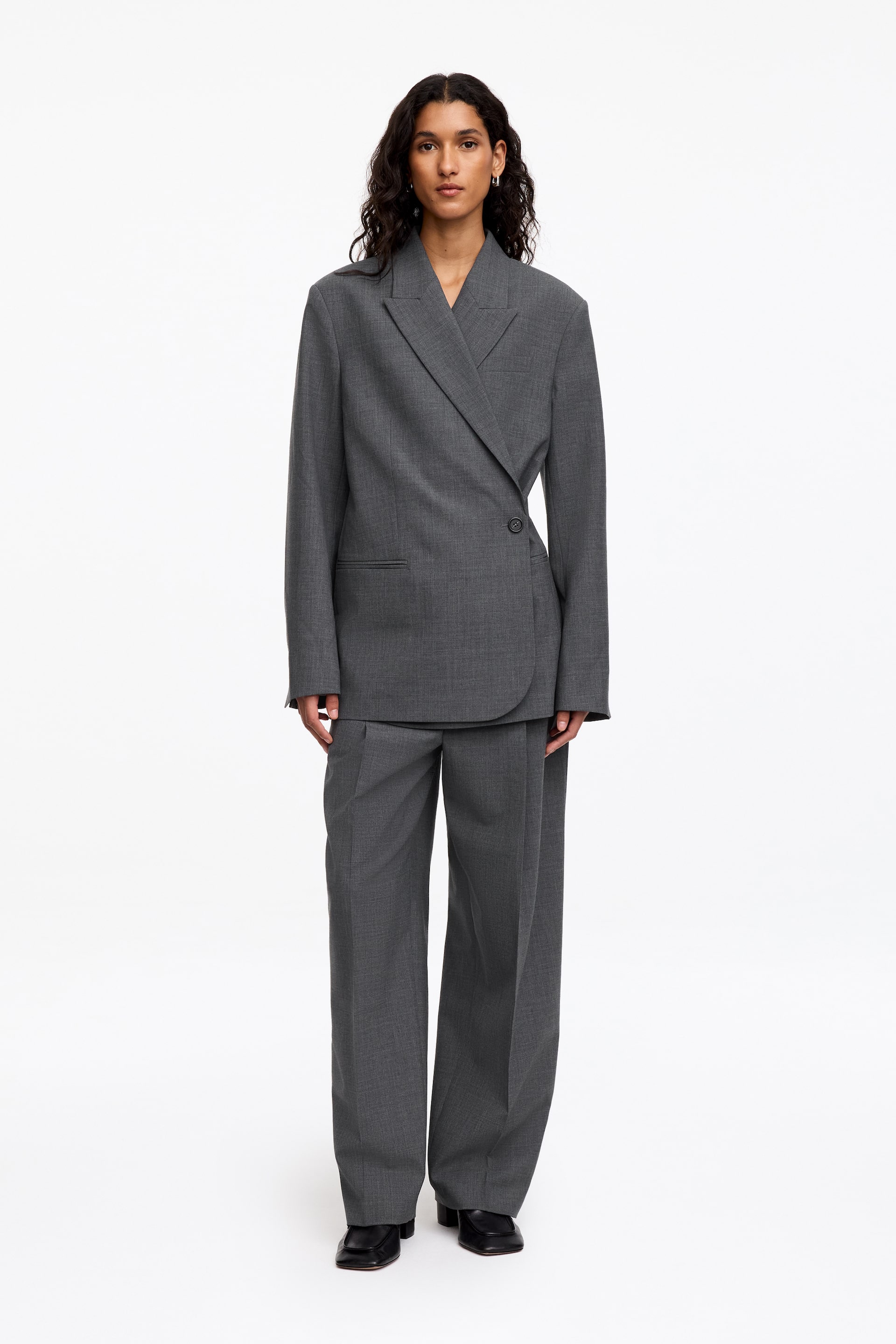 View larger image: The model is wearing a grey suit, comprising an oversized double-breasted blazer with notched lapels and structured shoulders, and matching wide-leg pleated trousers. The blazer is fastened with a single button, and the trousers drape over black shoes.