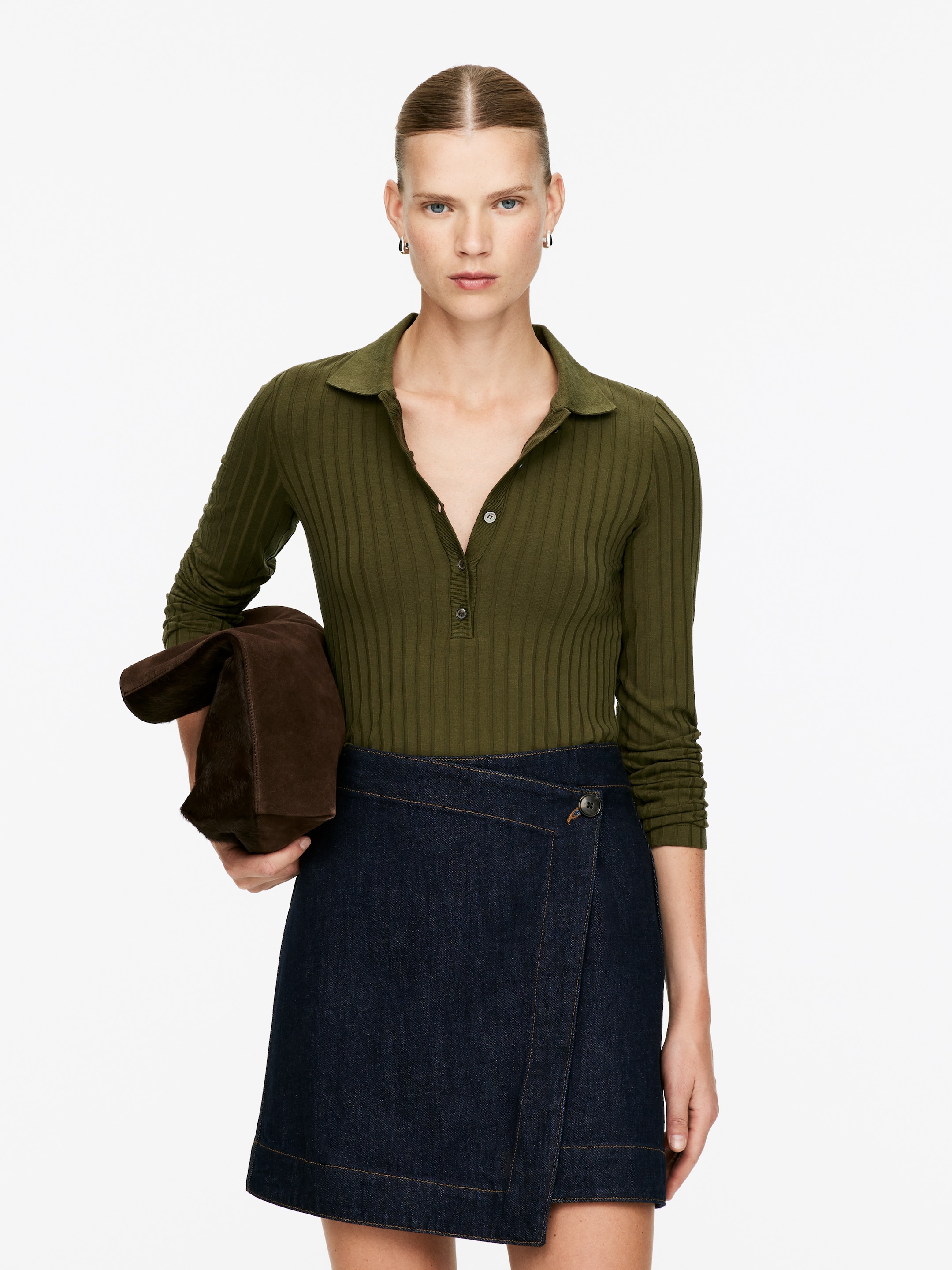 Größeres Bild ansehen: Ein olivgrünes, geripptes Langarm-Poloshirt mit Kragen und einer teilweisen Knopfleiste. Es wird mit einem dunkel gewaschenen Denim-Wickel-Minirock kombiniert, der mit einem sichtbaren Knopf geschlossen wird.